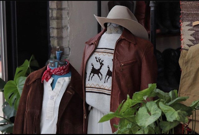Two mannequins dressed in leather jackets are seen situated between two plants.
