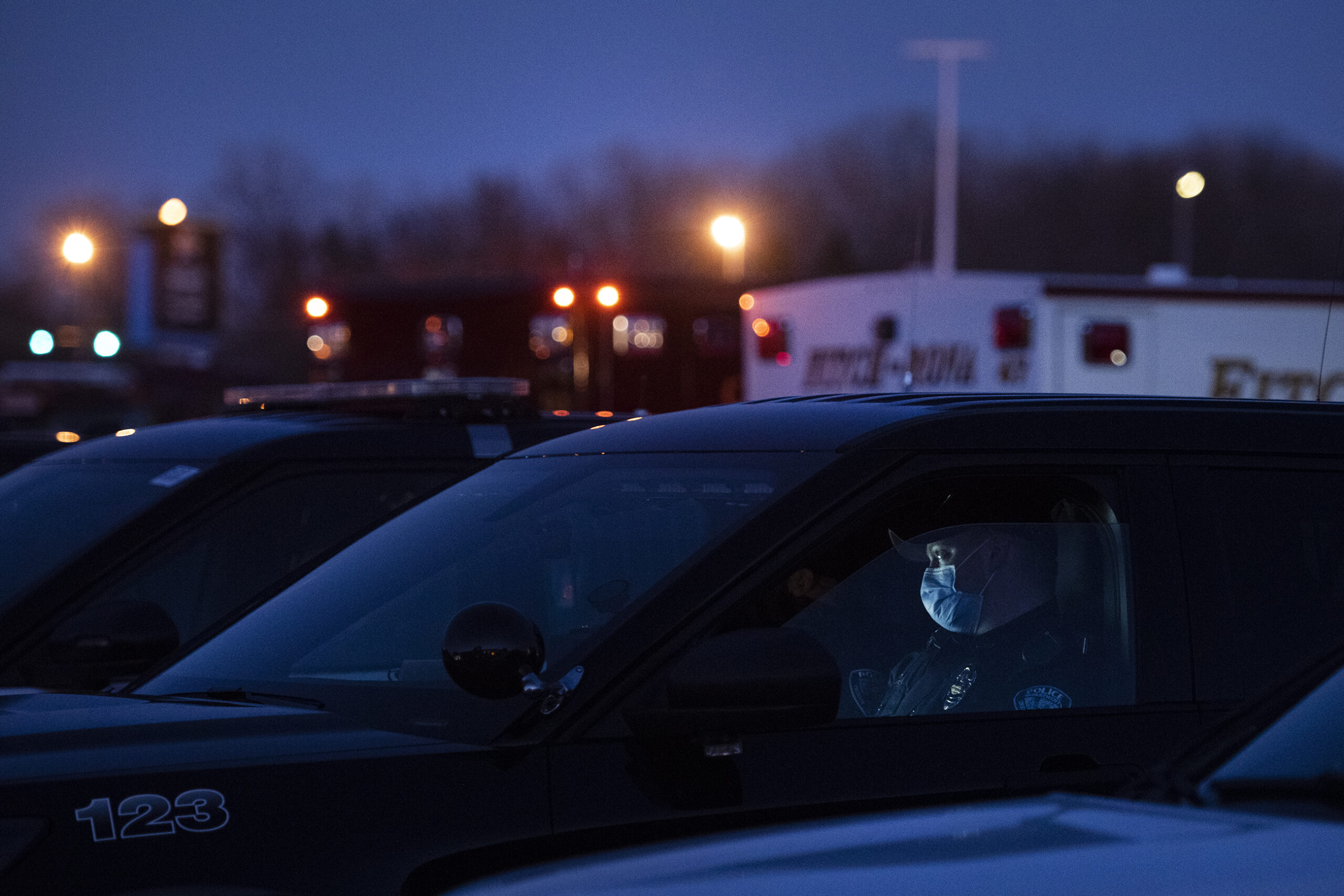 A police officer in a mask sits in a vehicle as the sky goes dark.