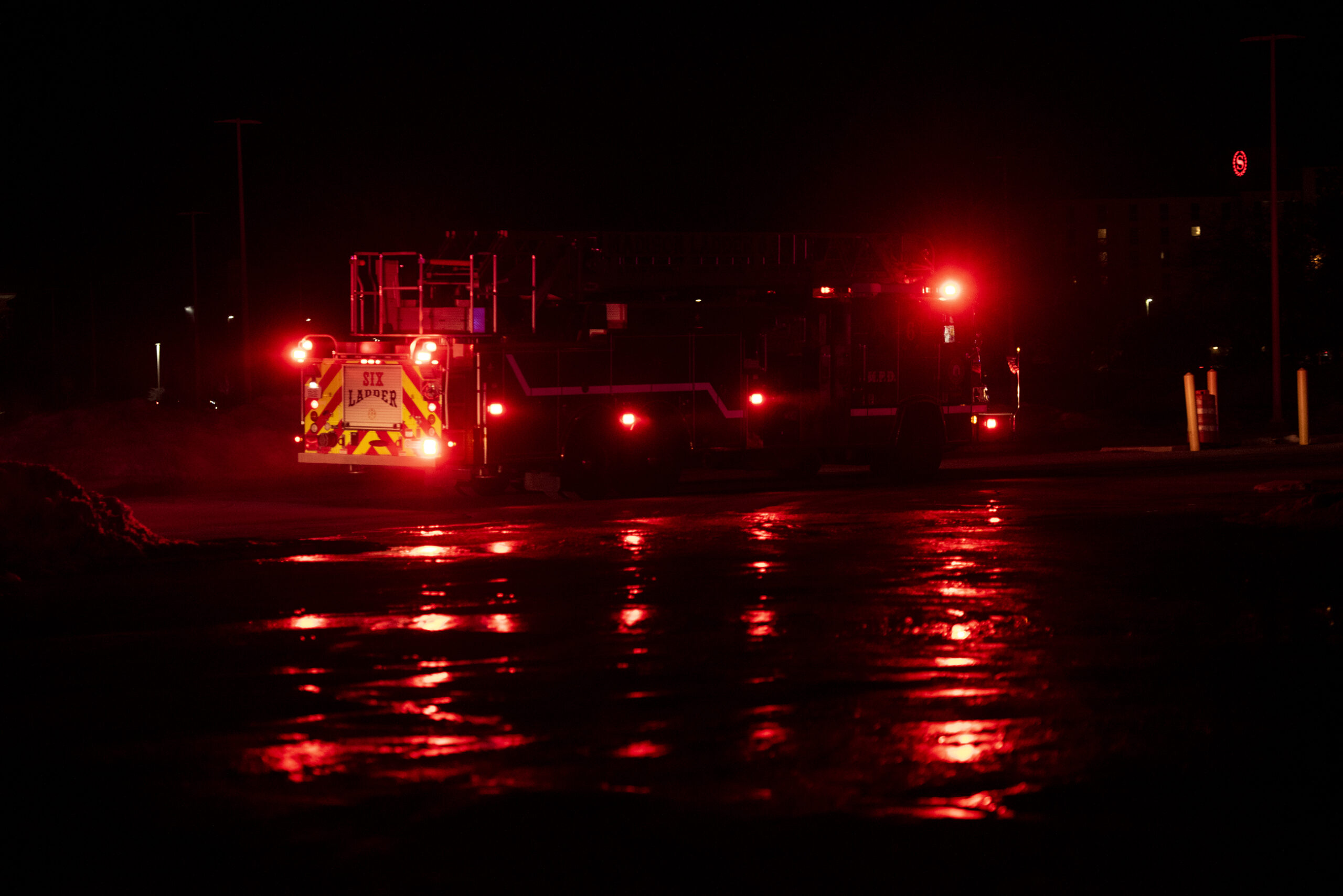 A dark night sky surrounds a firetruck with red lights on. Ice and water on the pavement glow with a reflection.