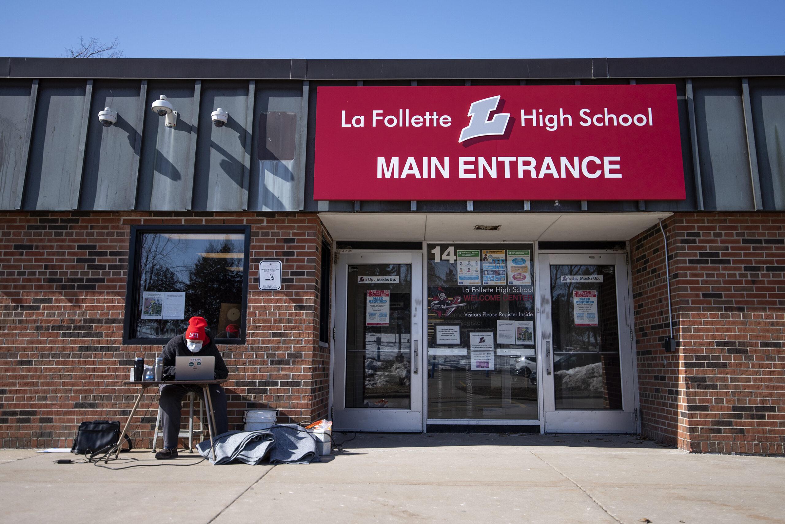 A red sign indicating the main entrance of La Follette High School is seen next to a teacher giving a lesson virtually outside.