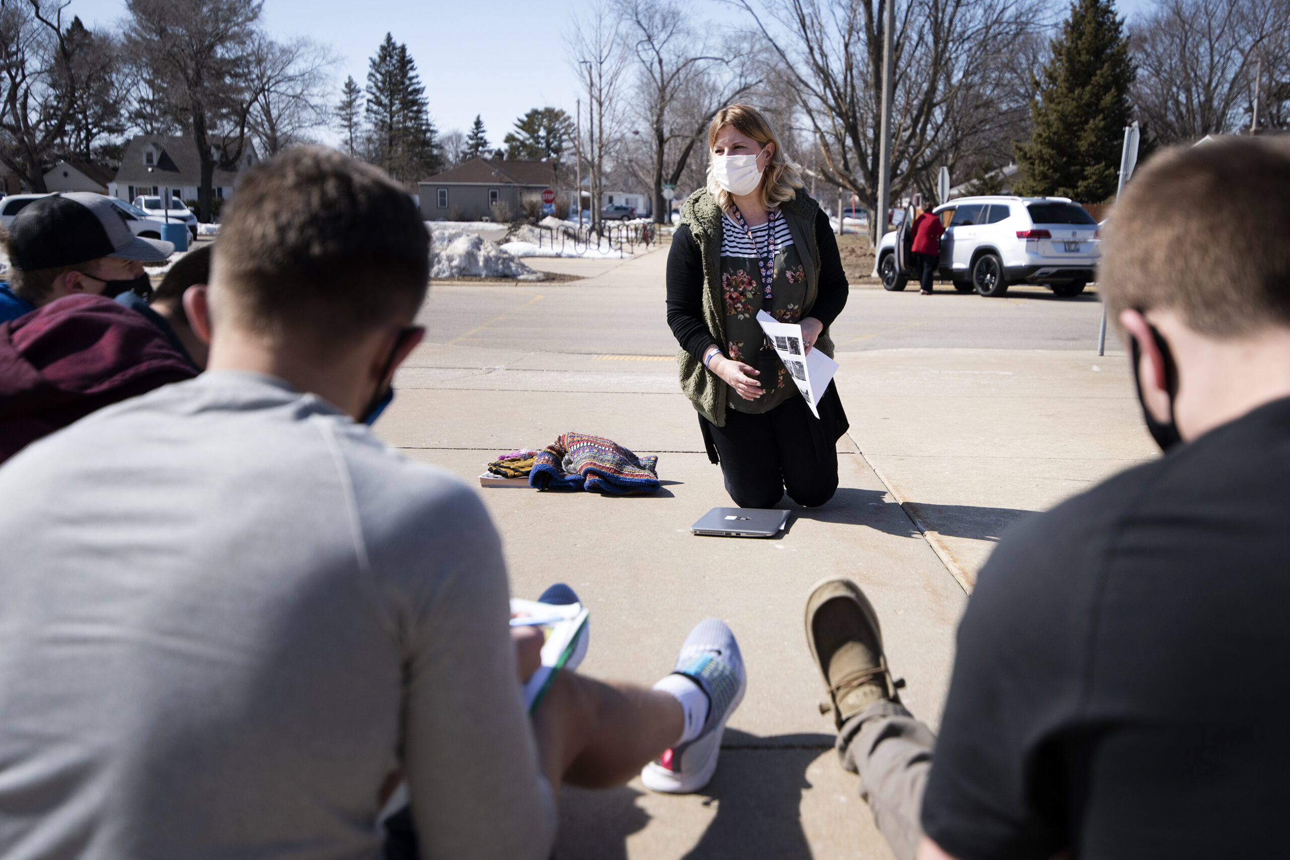 A teacher sits on the ground while wearing a face mask as a circle of students listen to her.