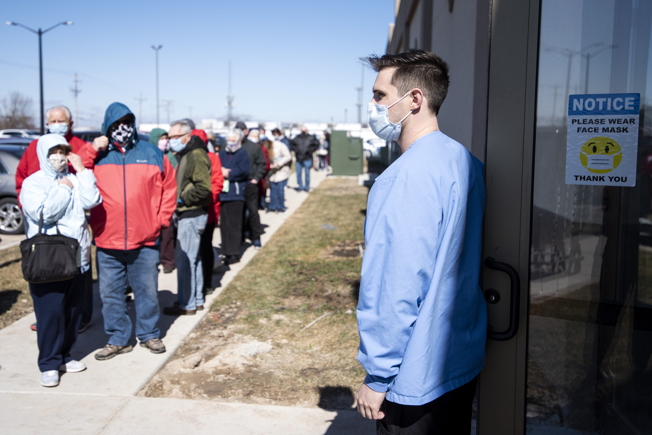 A line of people in jackets and face masks wait to enter a door held open by a medical worker.
