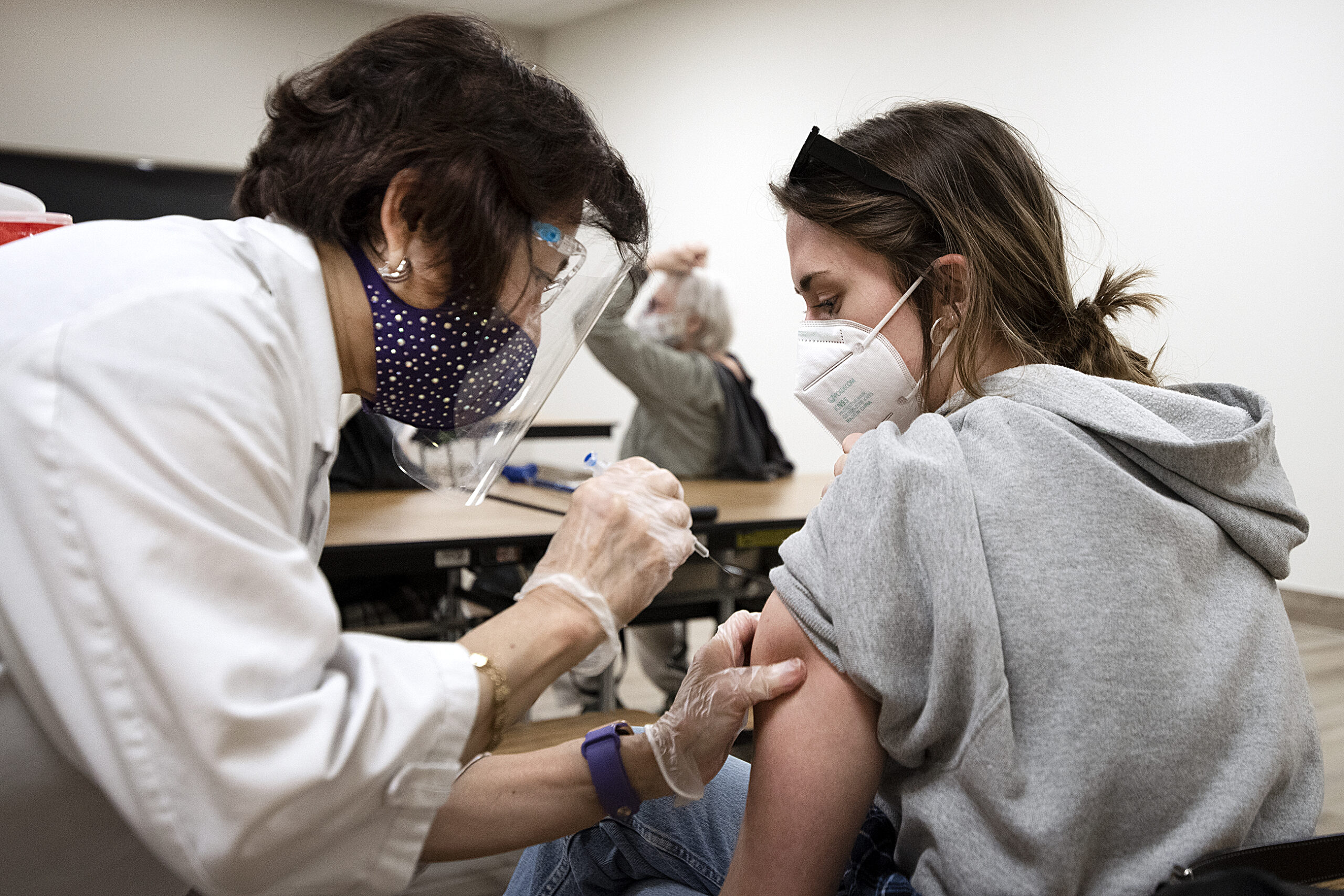 A woman in a face mask looks down at her arm as she receives a shot.