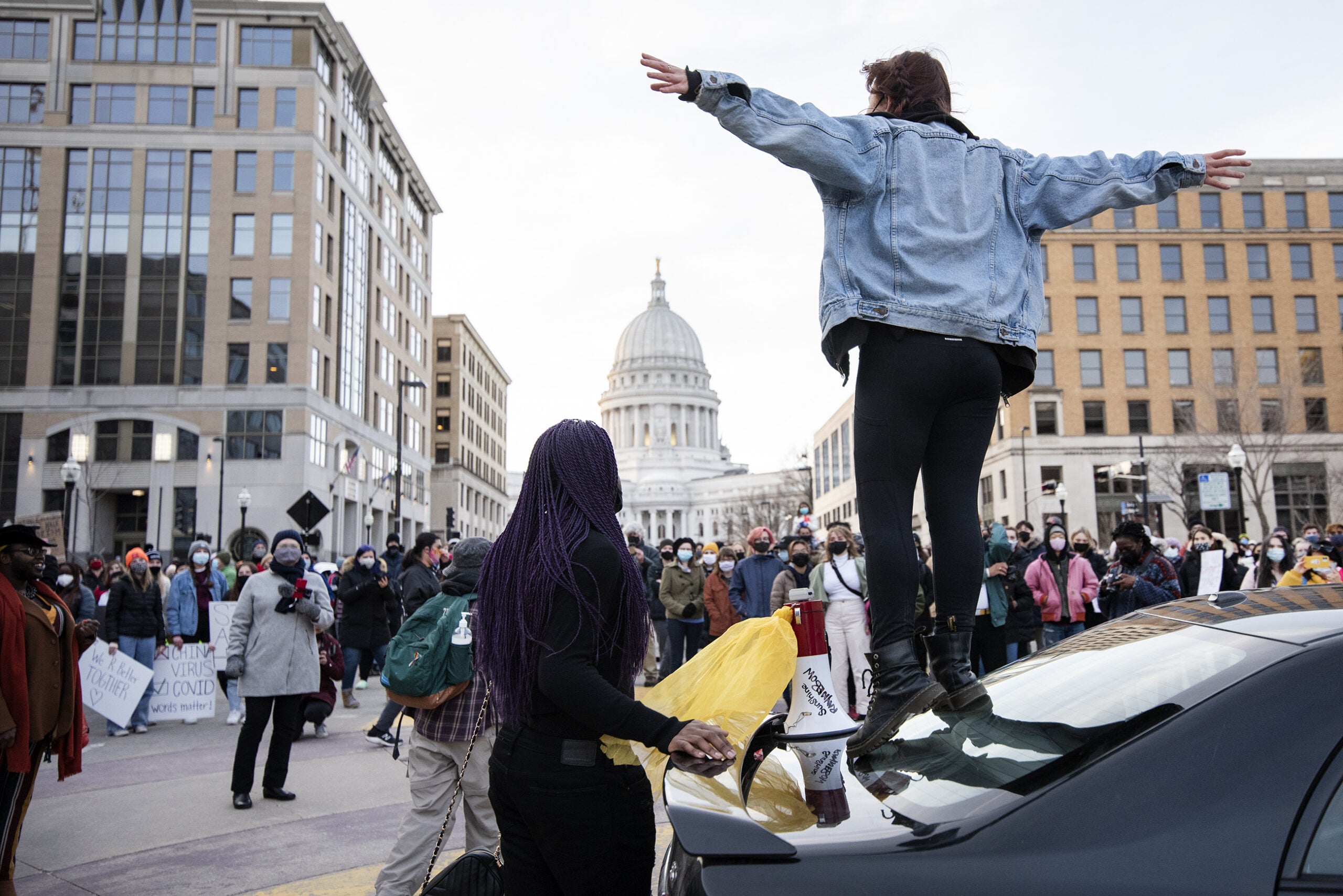 A woman in a jean jacket spreads her arms as she speaks to a crowd of masked protesters.