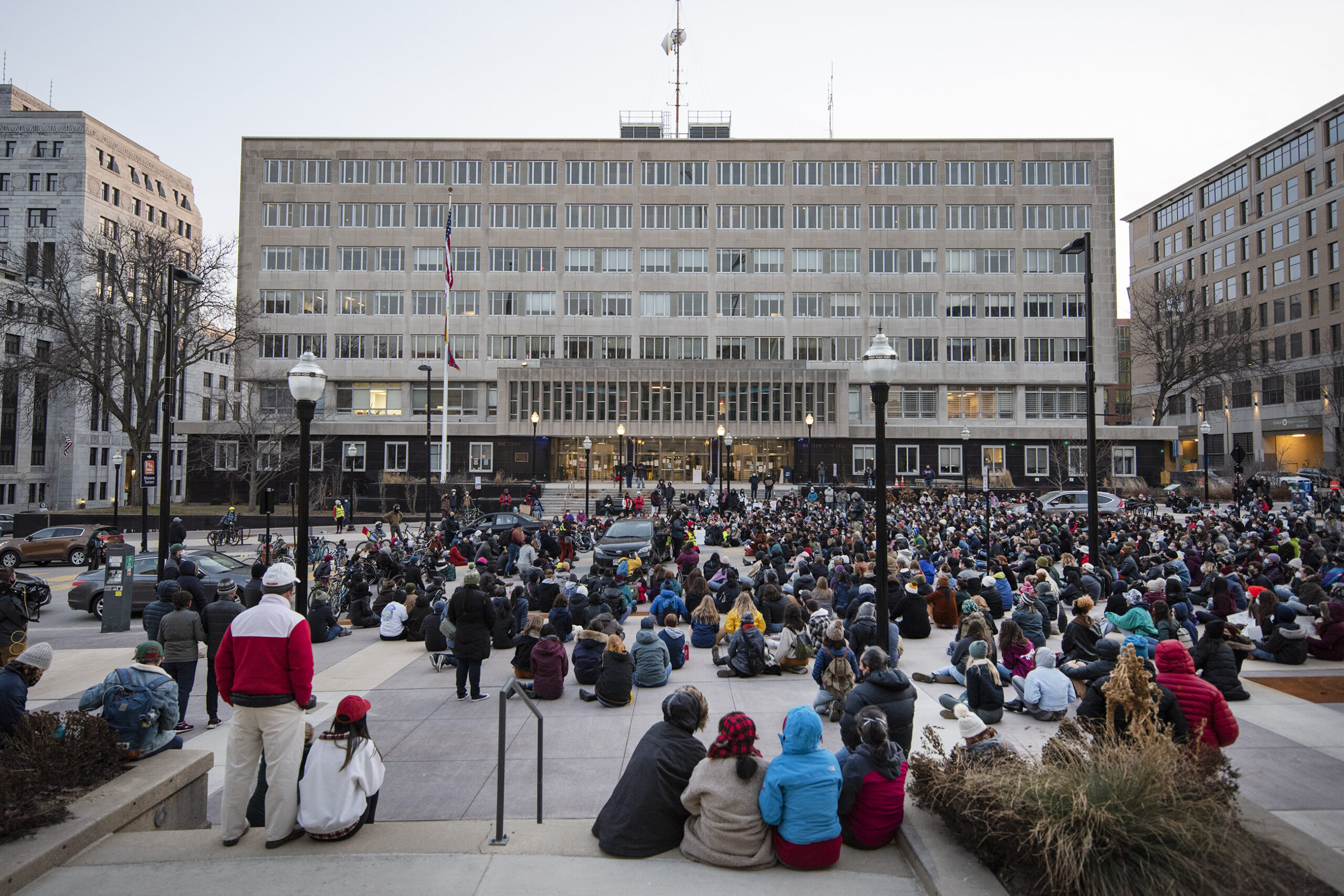 A crowd of people sit on the road as they listen to speakers.