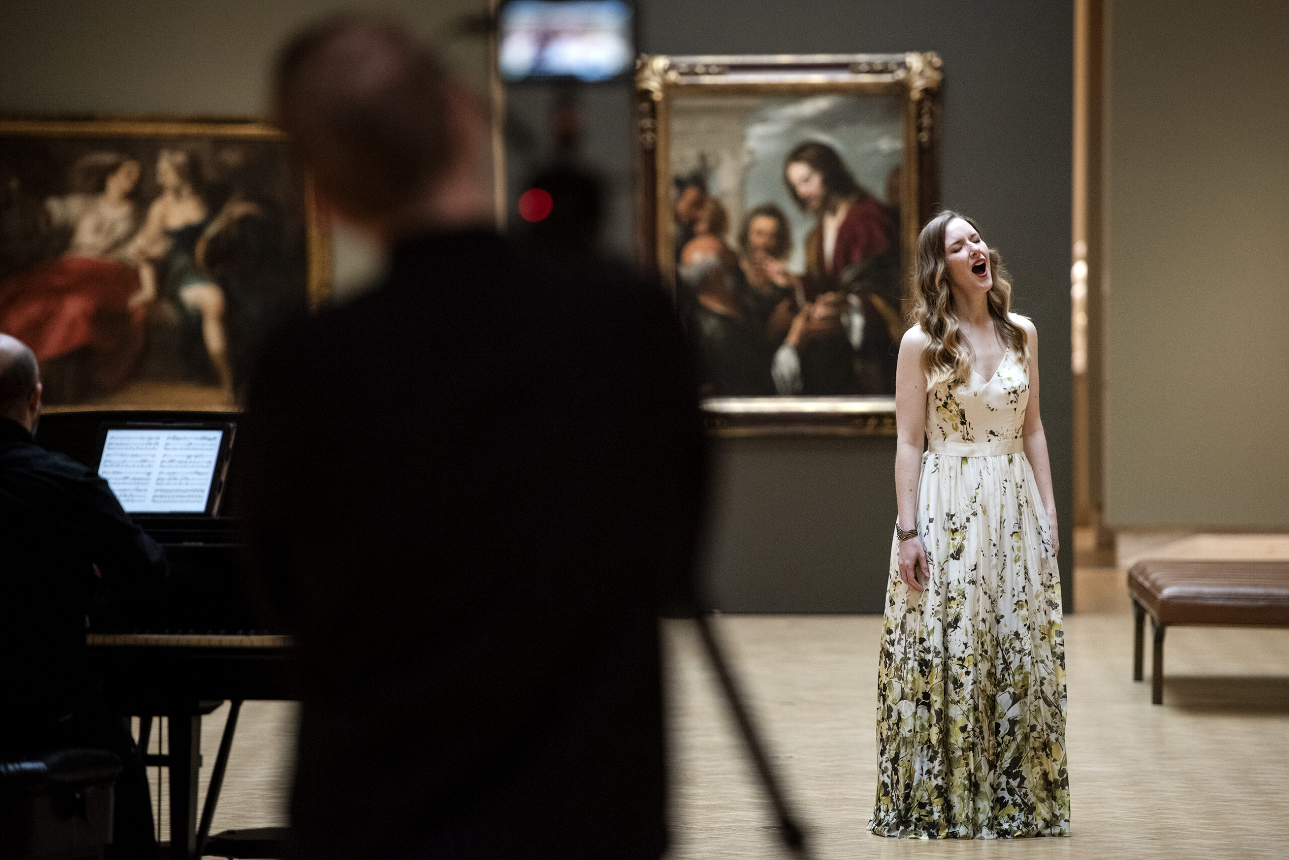 A singer in a long white floral gown tilts her head back as she belts a note surrounded by paintings in a museum.