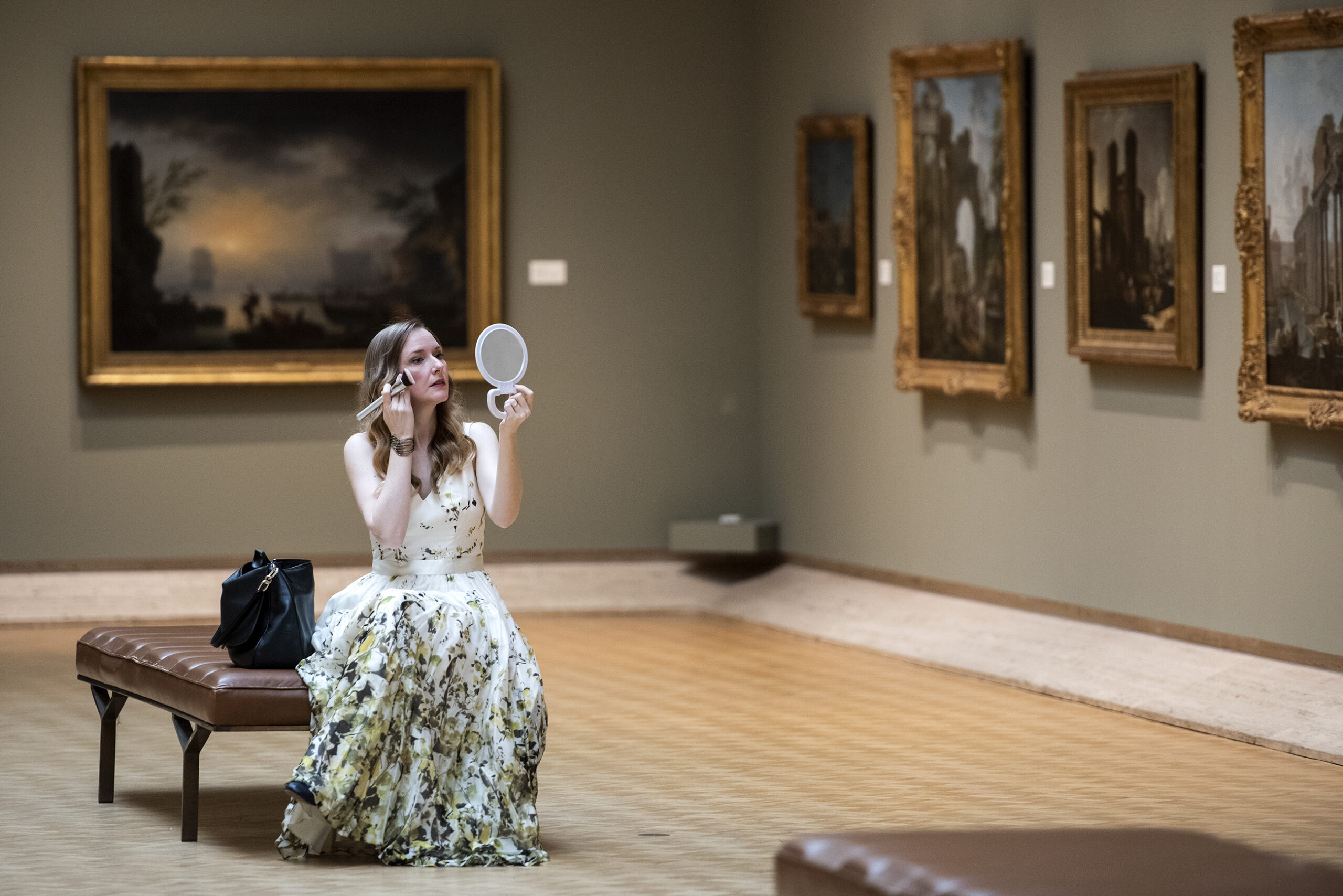 A woman in a white gown sits on a bench as she holds a makeup brush and a mirror.