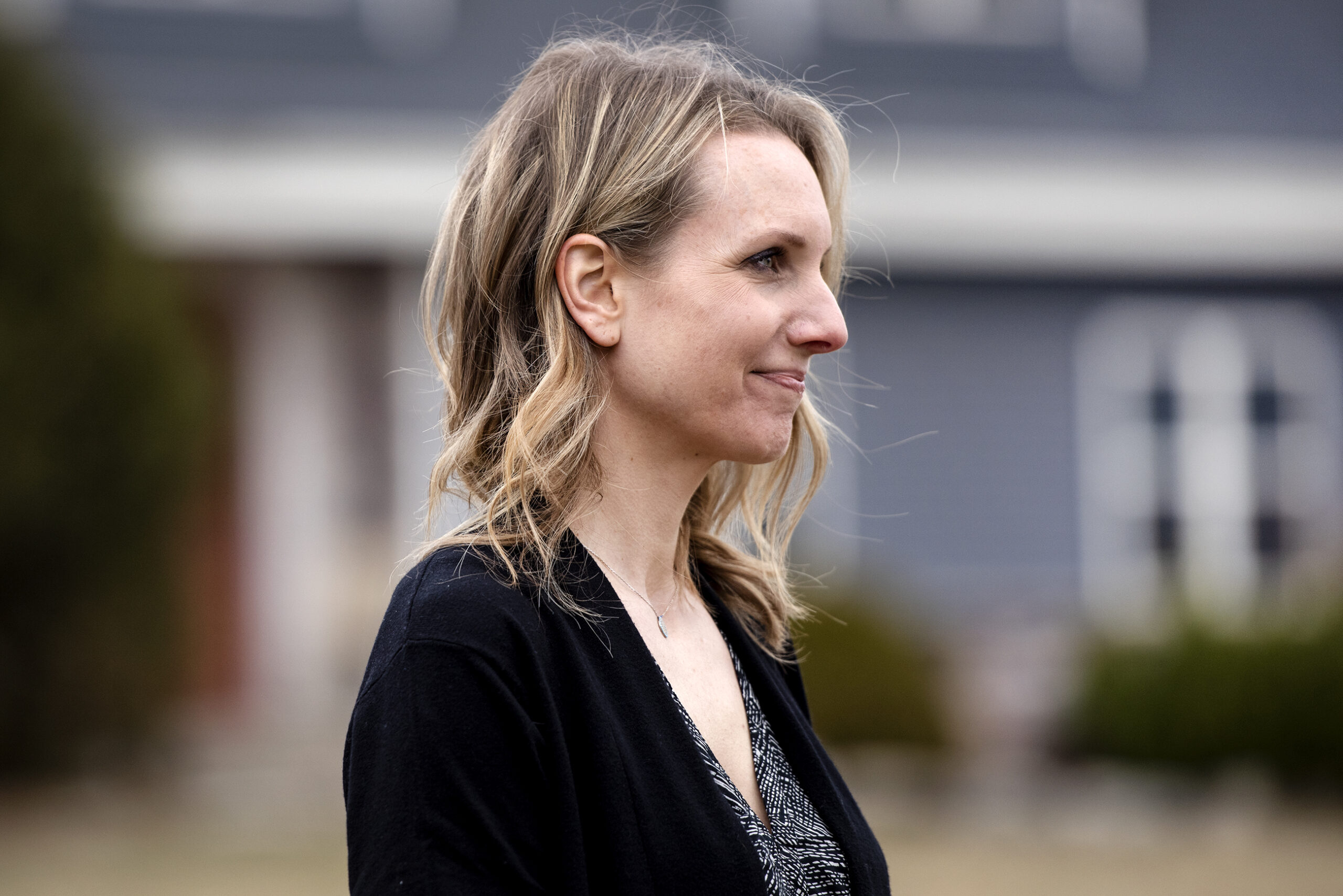 A woman smiles slightly as she looks to the side while standing in front of a blue house.