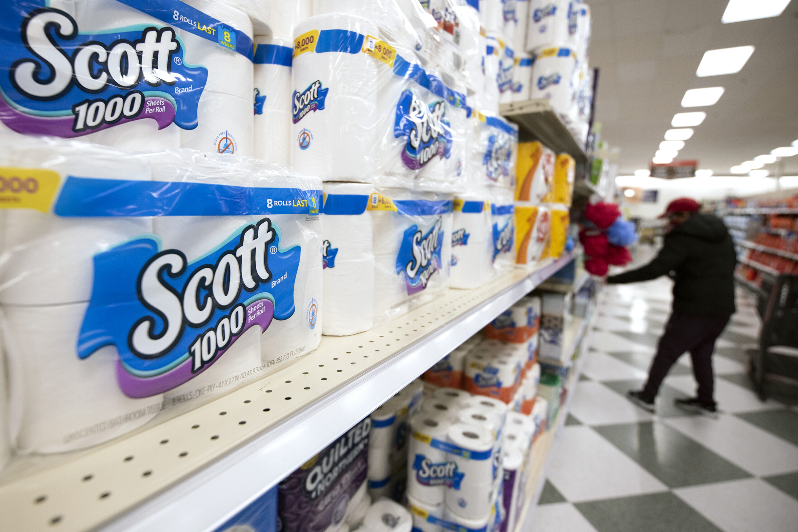 Scott toilet paper is on a shelf. A customer can be seen in the background reaching for an item.