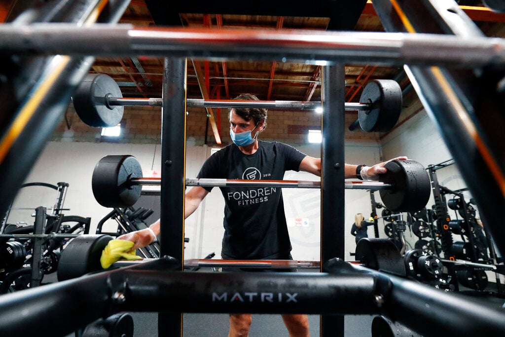 A man wipes off a pre-loaded barbell with a sanitizing cloth on the floor of a gym. Other barbell and racks can be seen in the background.