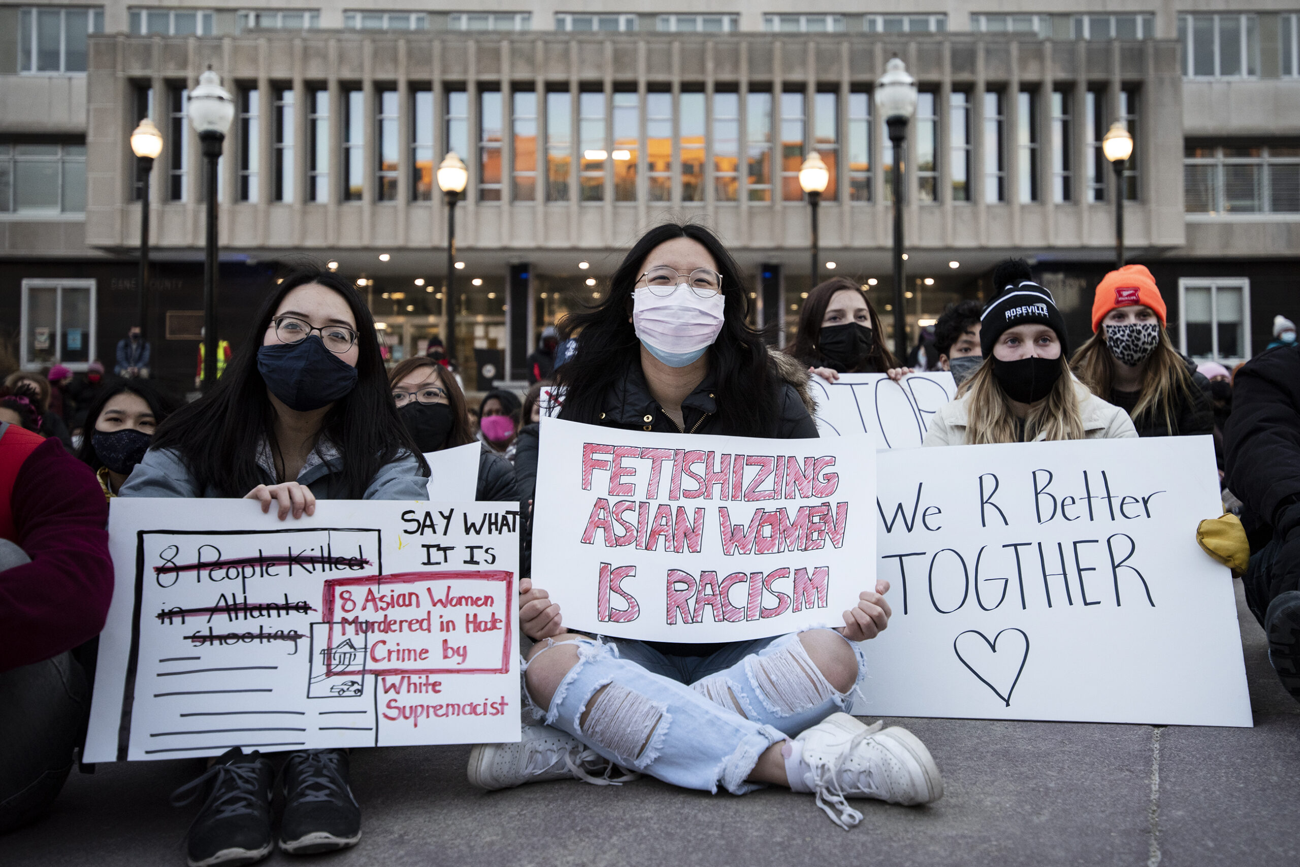 Three women sit prominently at the front of a crowd wearing face masks and displaying signs in support of Asian women.
