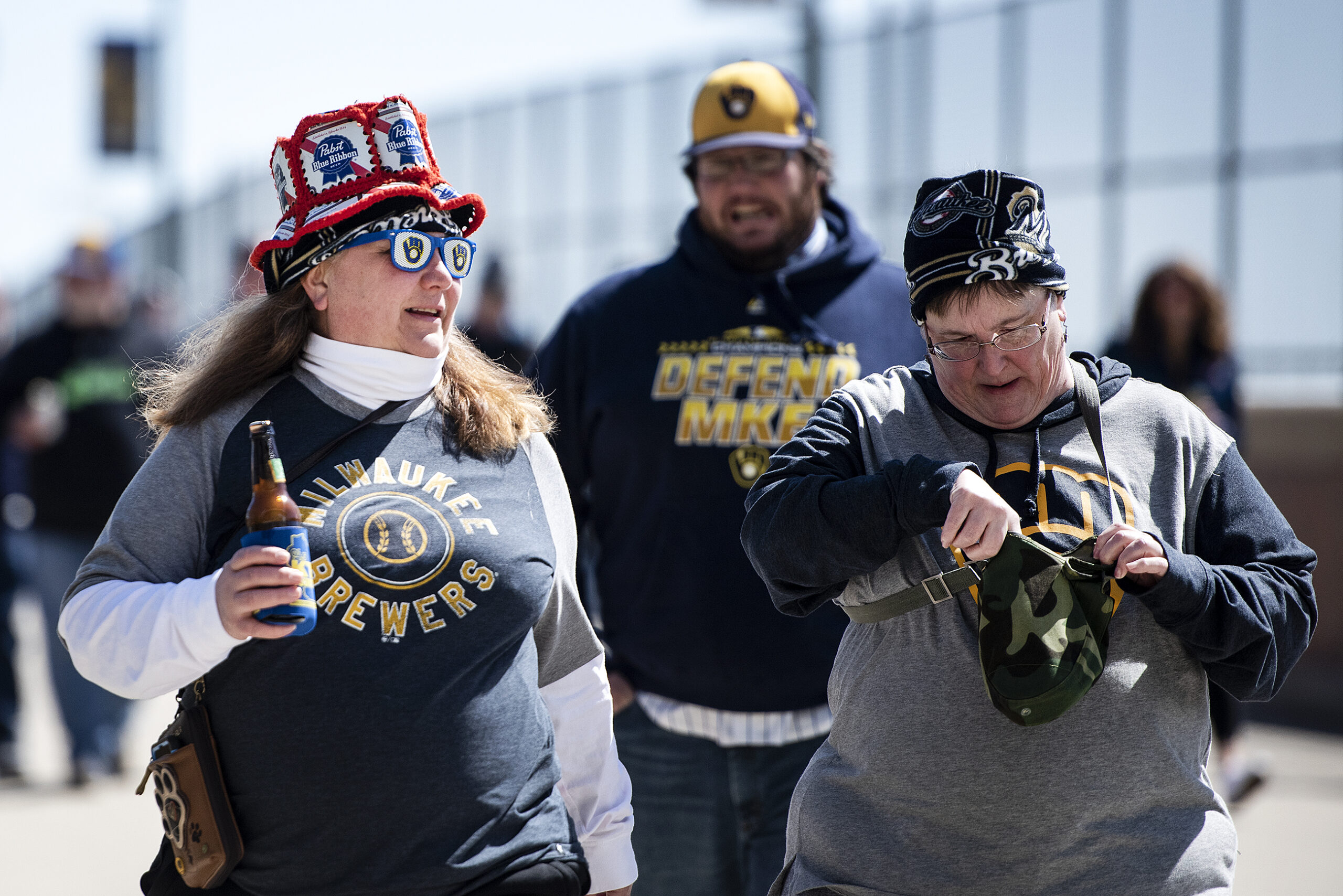 A fan wears a glasses with the Brewers logo, a hat made of PBR boxes and a Brewers shirt as she holds a beer walking into the stadium.