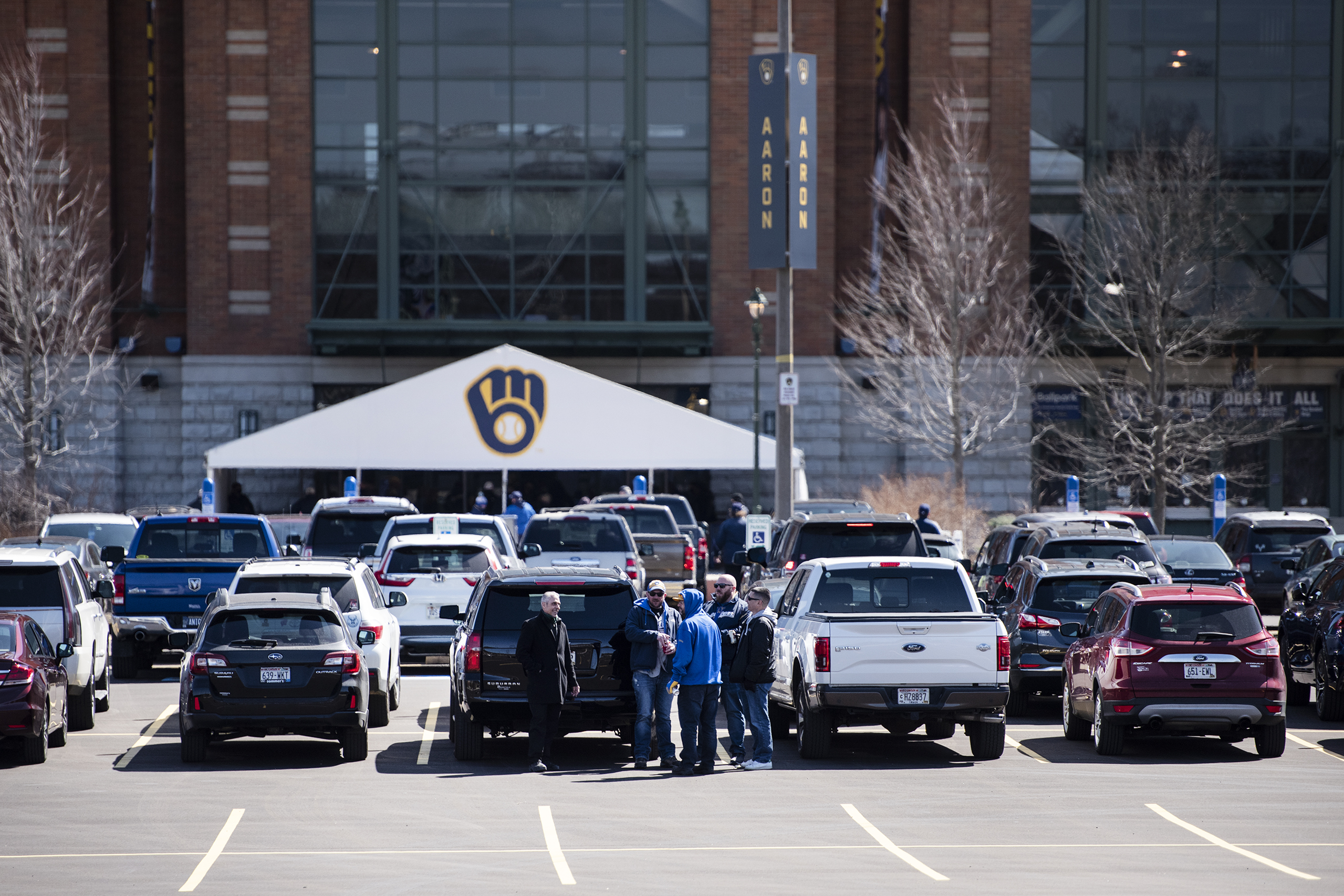 Cars are parked in the parking lot in front of American Family Field. A small group of men gather before entering.