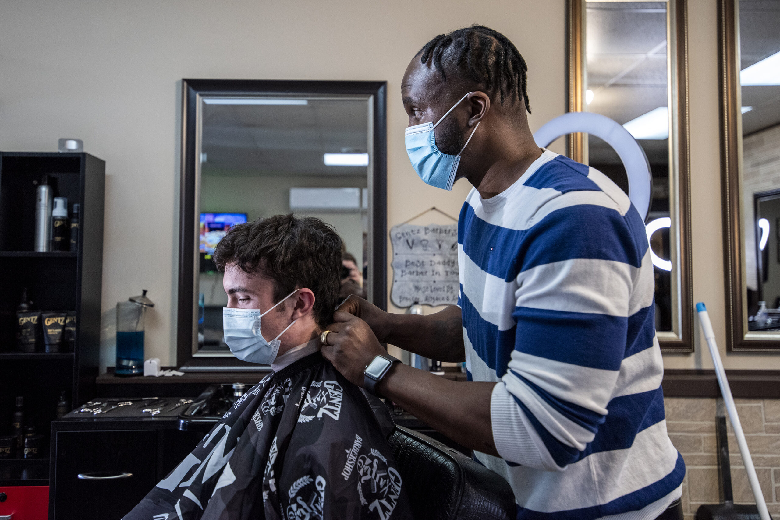 A barber in a face mask ties a smock onto a client in his chair.
