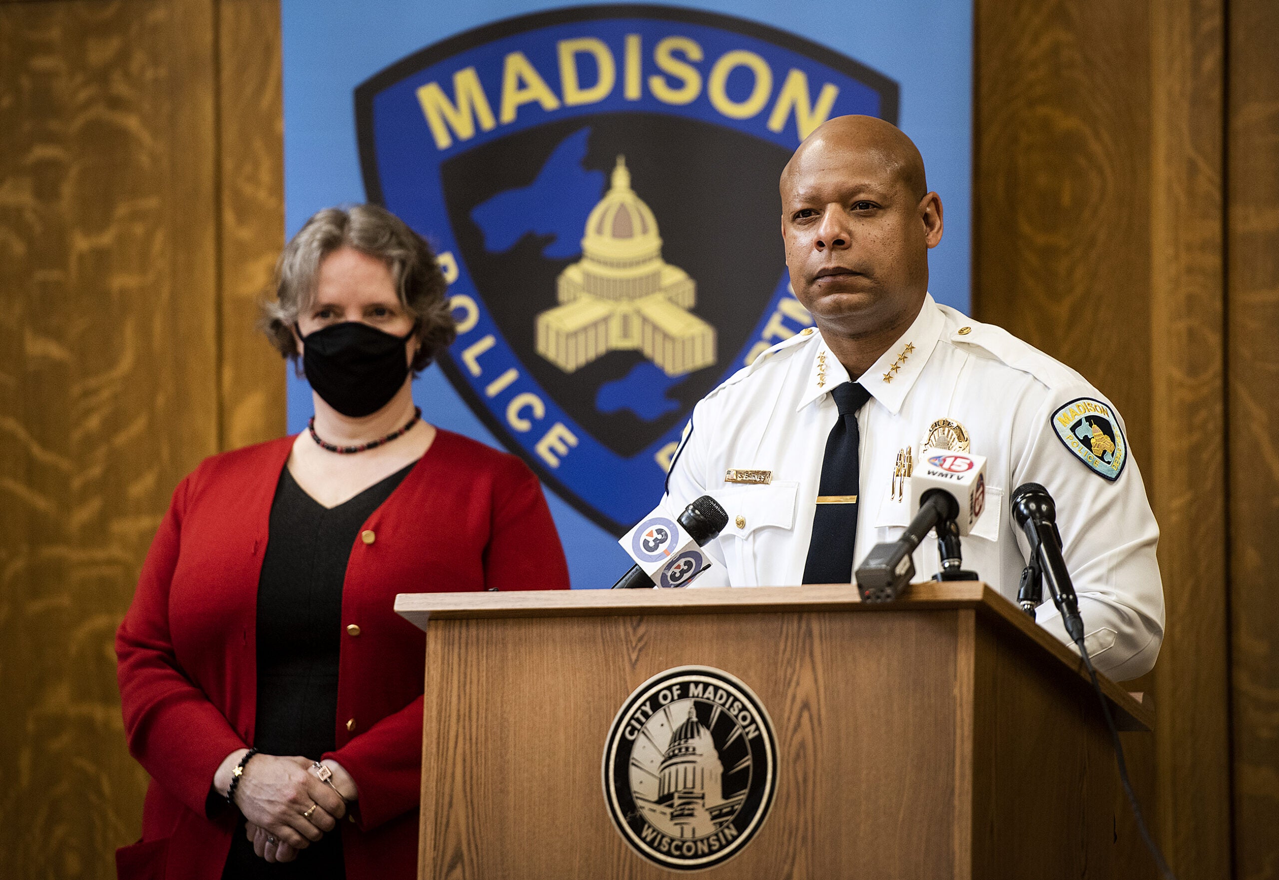 A man stands at a podium in a police uniform. Mayor Rhodes-Conway stands off to the side.