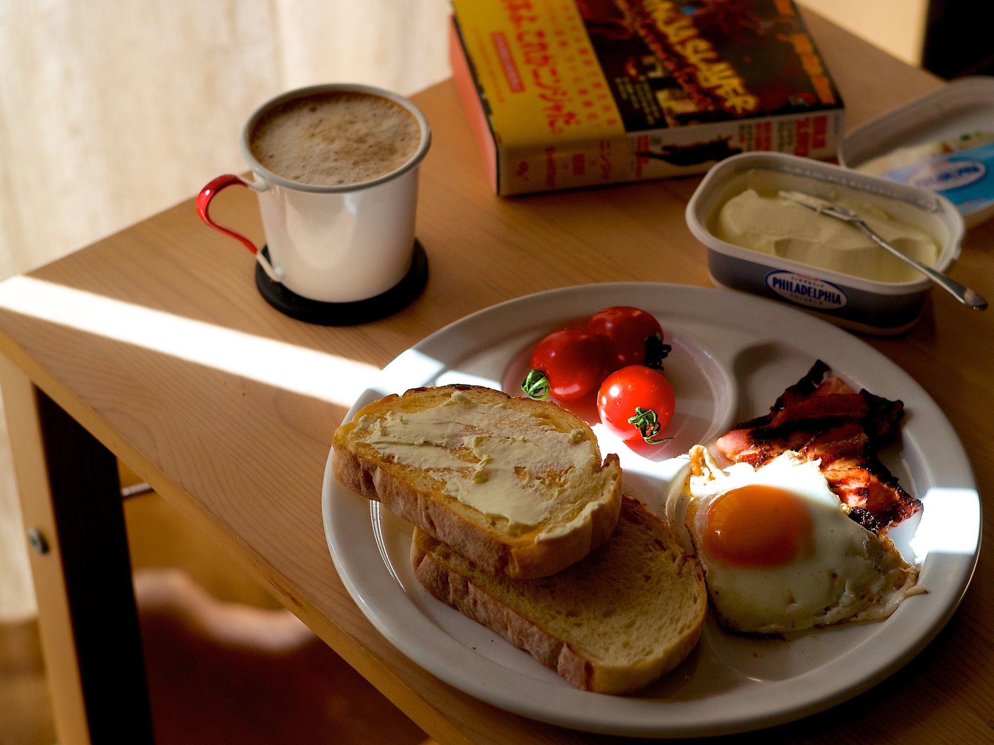 A table with a plate full of eggs and toast, a mug full of hot beverage and a book sit in a sunbeam.