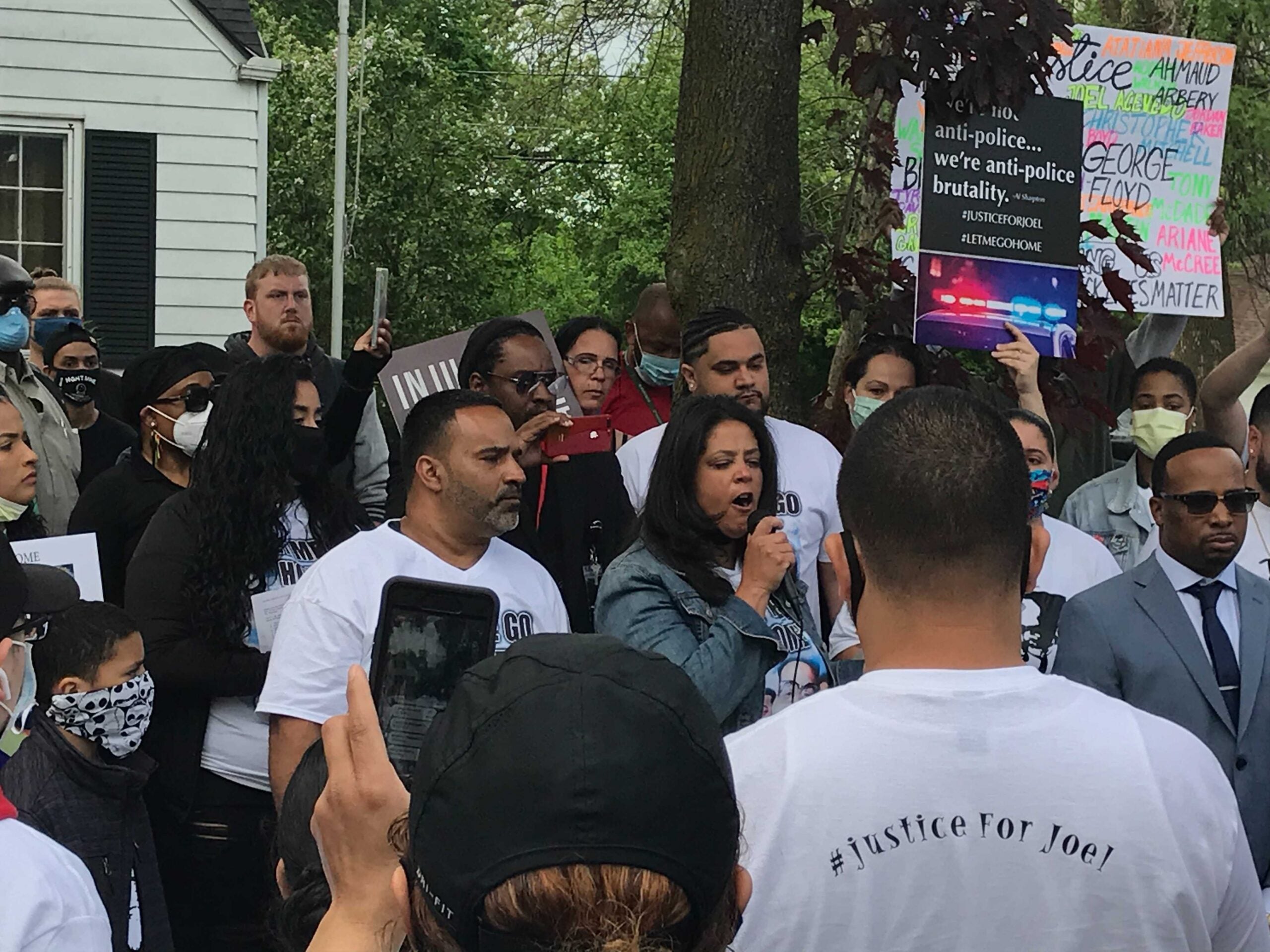 A woman with a microphone speaks to a crowd holding signs outside a home