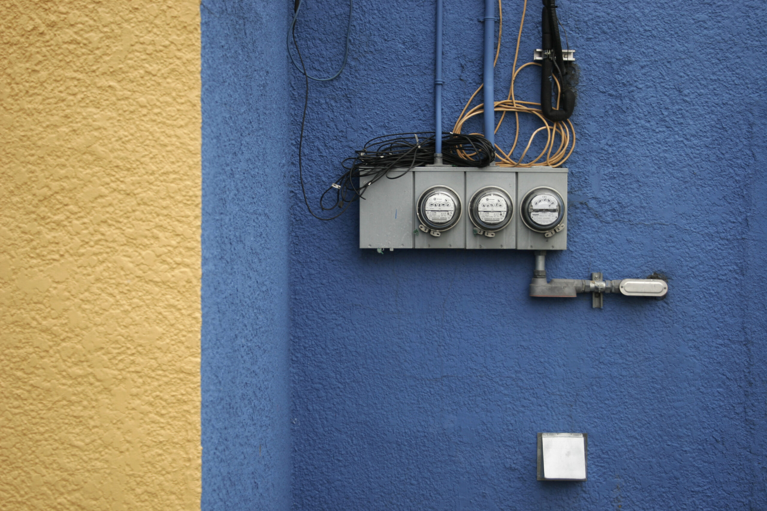 Yellow and blue stucco wall decorated with a scattershot corsage of electric meters, conduit and wiring.