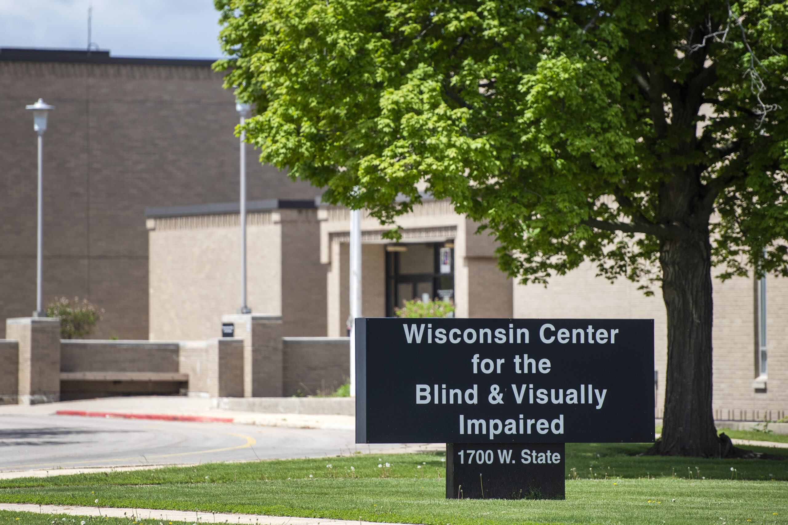 A sign says "Wisconsin Center for the Blind and Visually Impaired" under a tree with green leaves in front of a brick building.