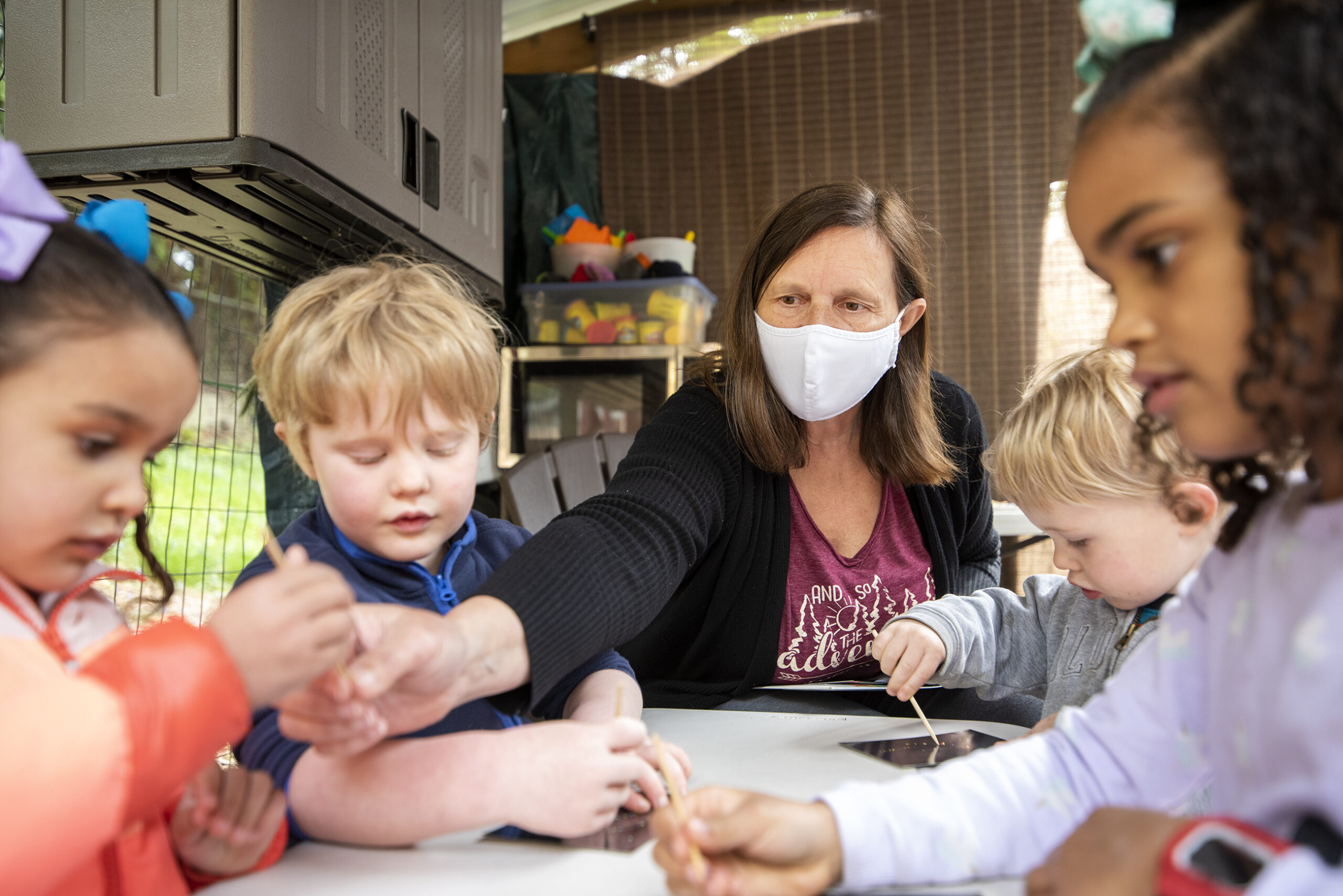 Four children sit at a small table. A woman in a face mask hands out craft tools.