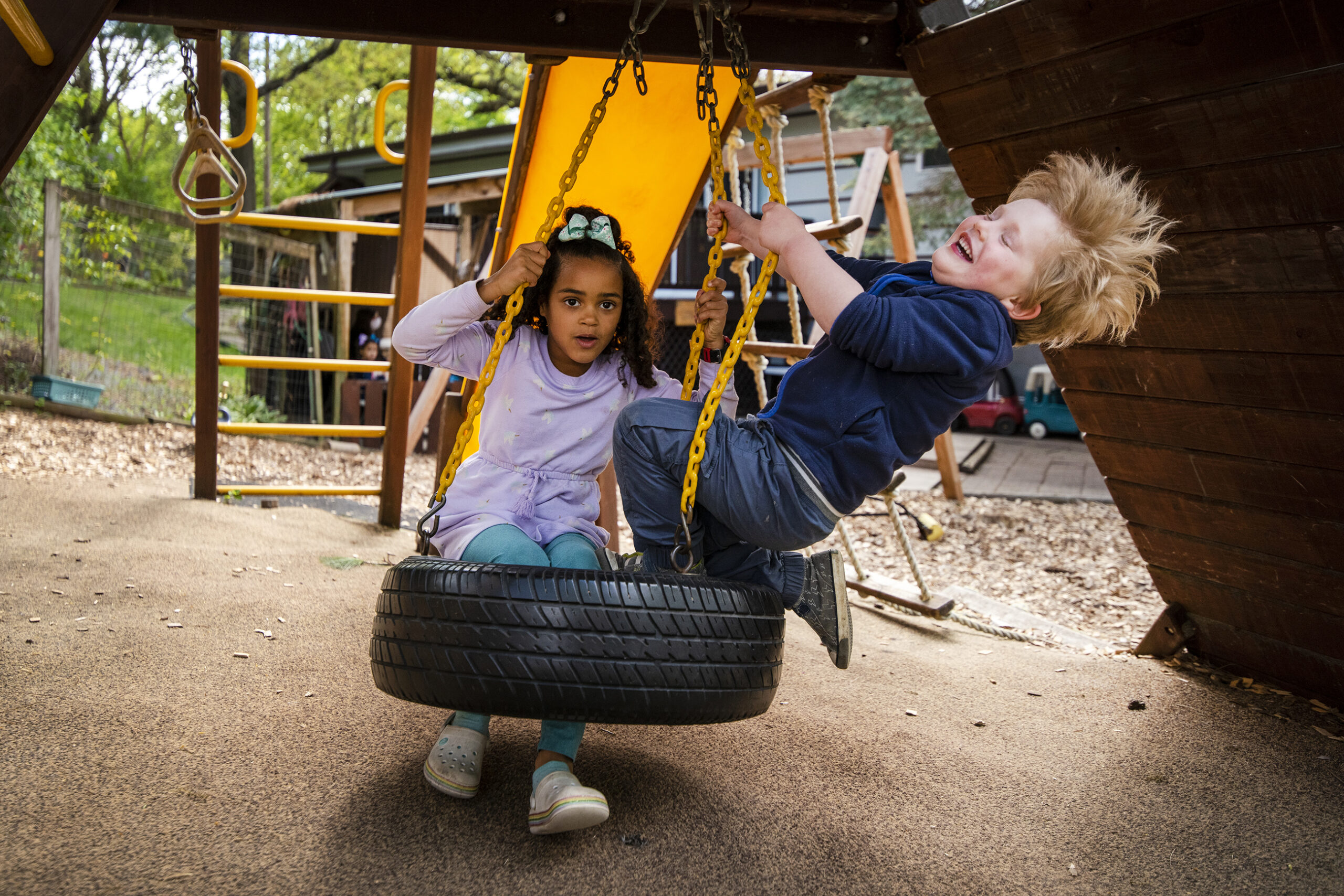 Two children swing on a tire swing. A child leans back as his blonde hair fans out behind him.