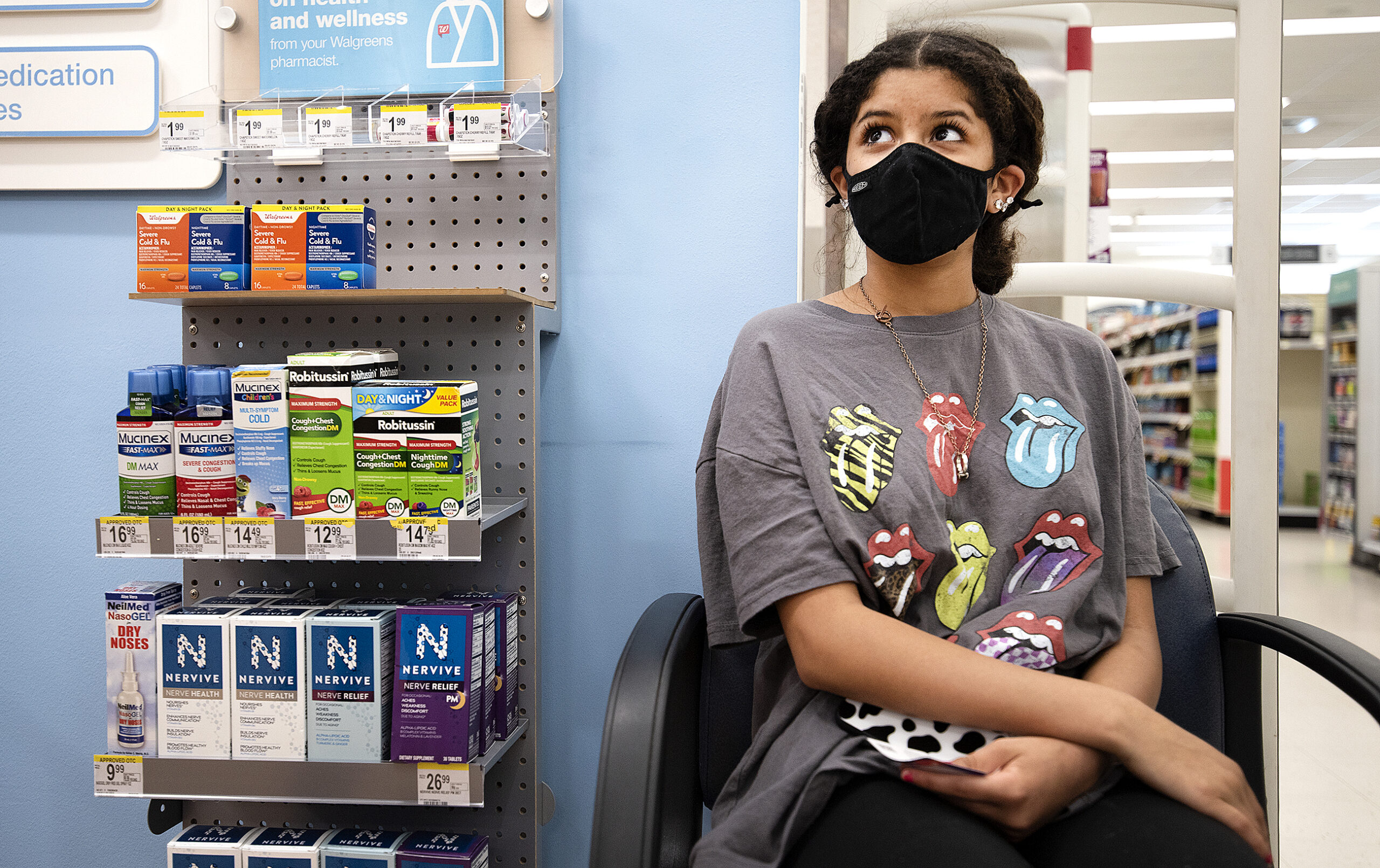 A girl in a t-shirt and face mask sits near products in a pharmacy.