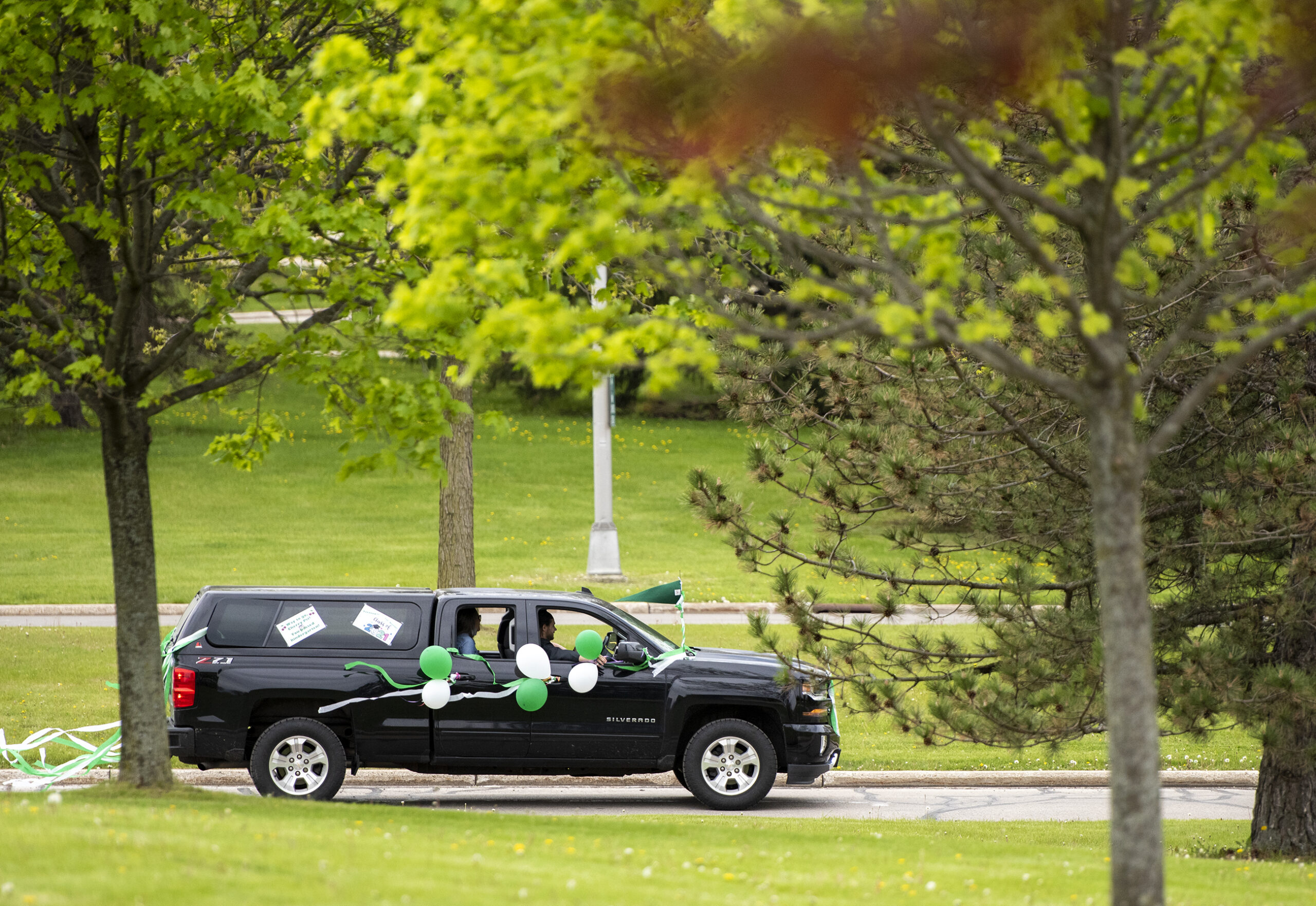 A black SUV with green and white balloons attached is seen in between green trees.