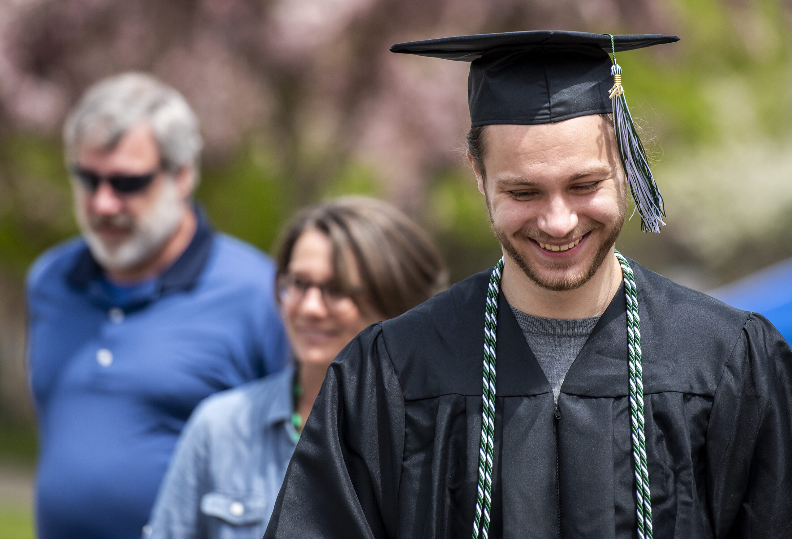 A graduate in robes and a cap looks down as he smiles. He's followed by family members behind him.