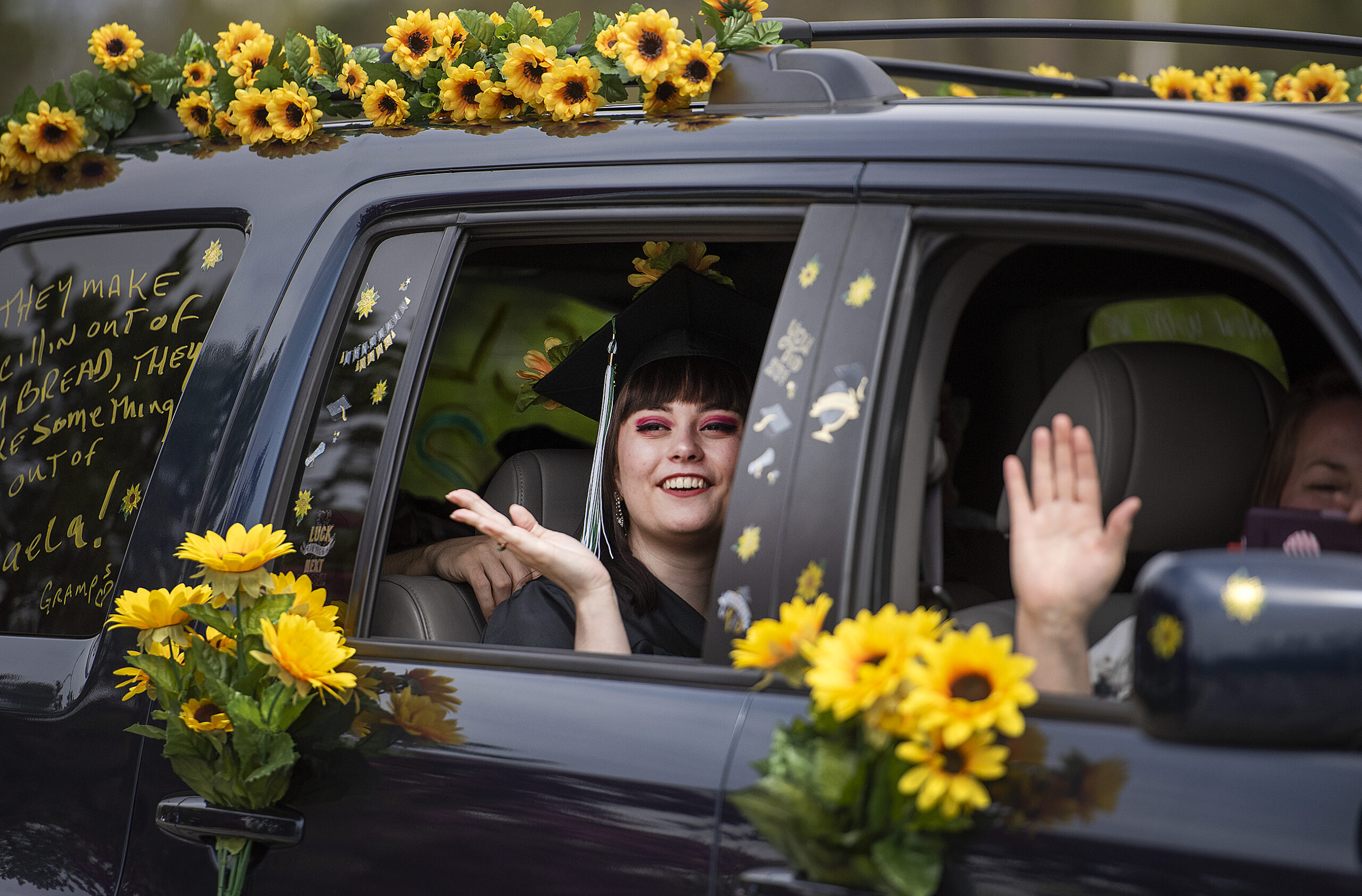 A graduate waves from inside of a vehicle decorated with yellow sunflowers.