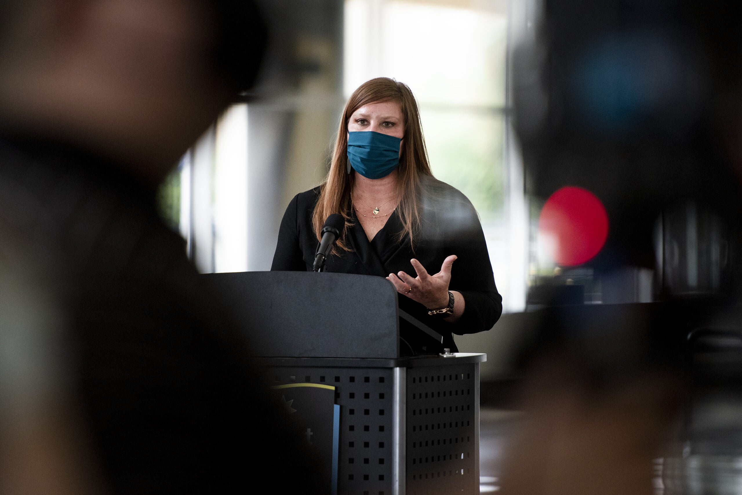 A woman is seen past the lens of a tv camera as she speaks at a press conference.