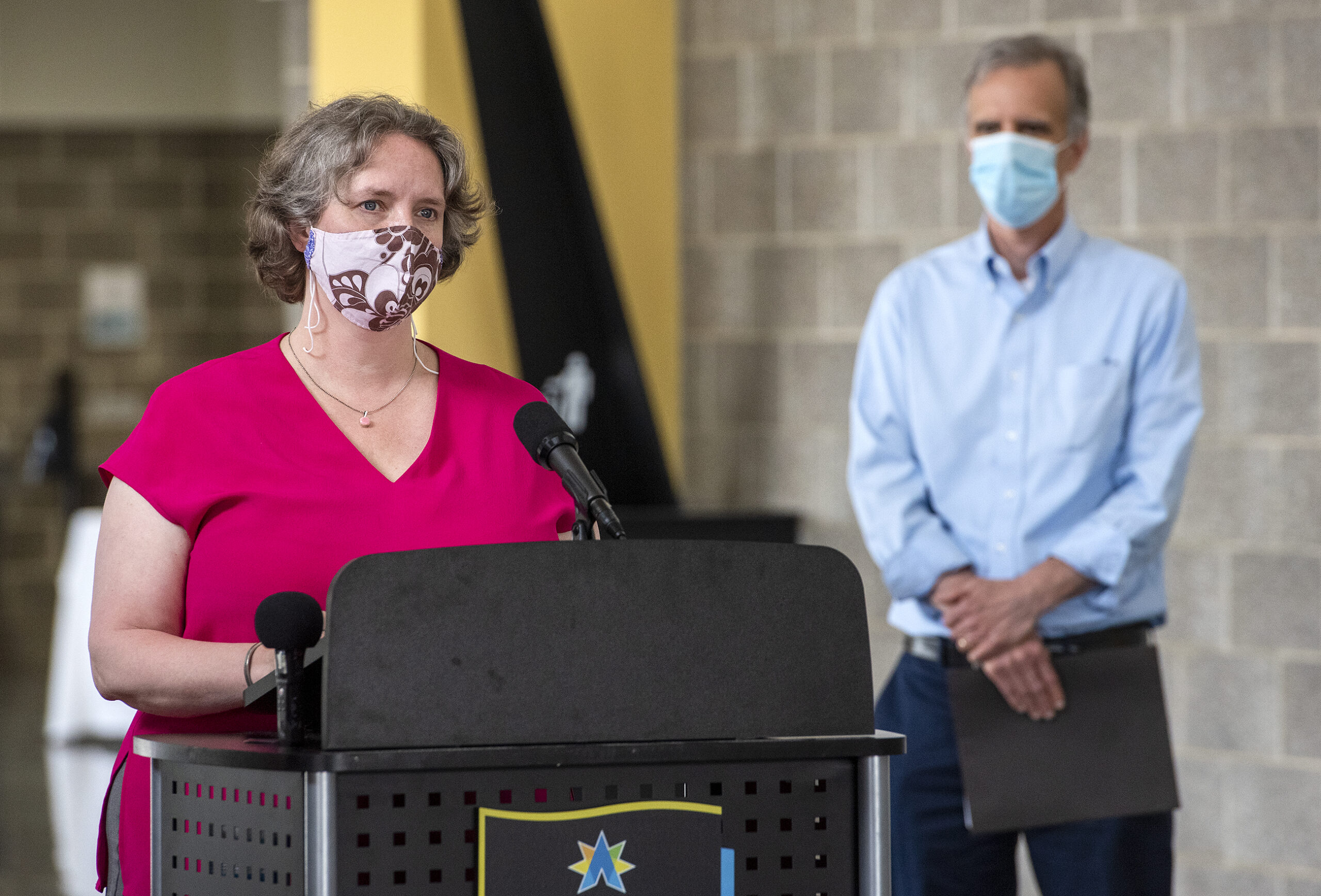 Satya Rhodes-Conway wears a mask while speaking at a podium. Joe Parisi stands off to the side, also in a face mask.