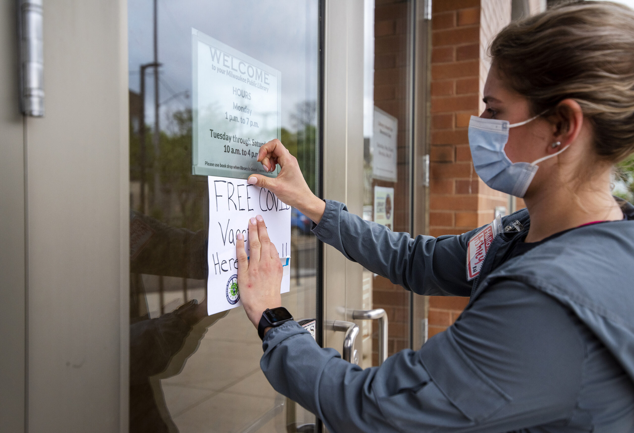 A nurse hangs a sign on a glass door that says "Free COVID-19 Vaccines Here"