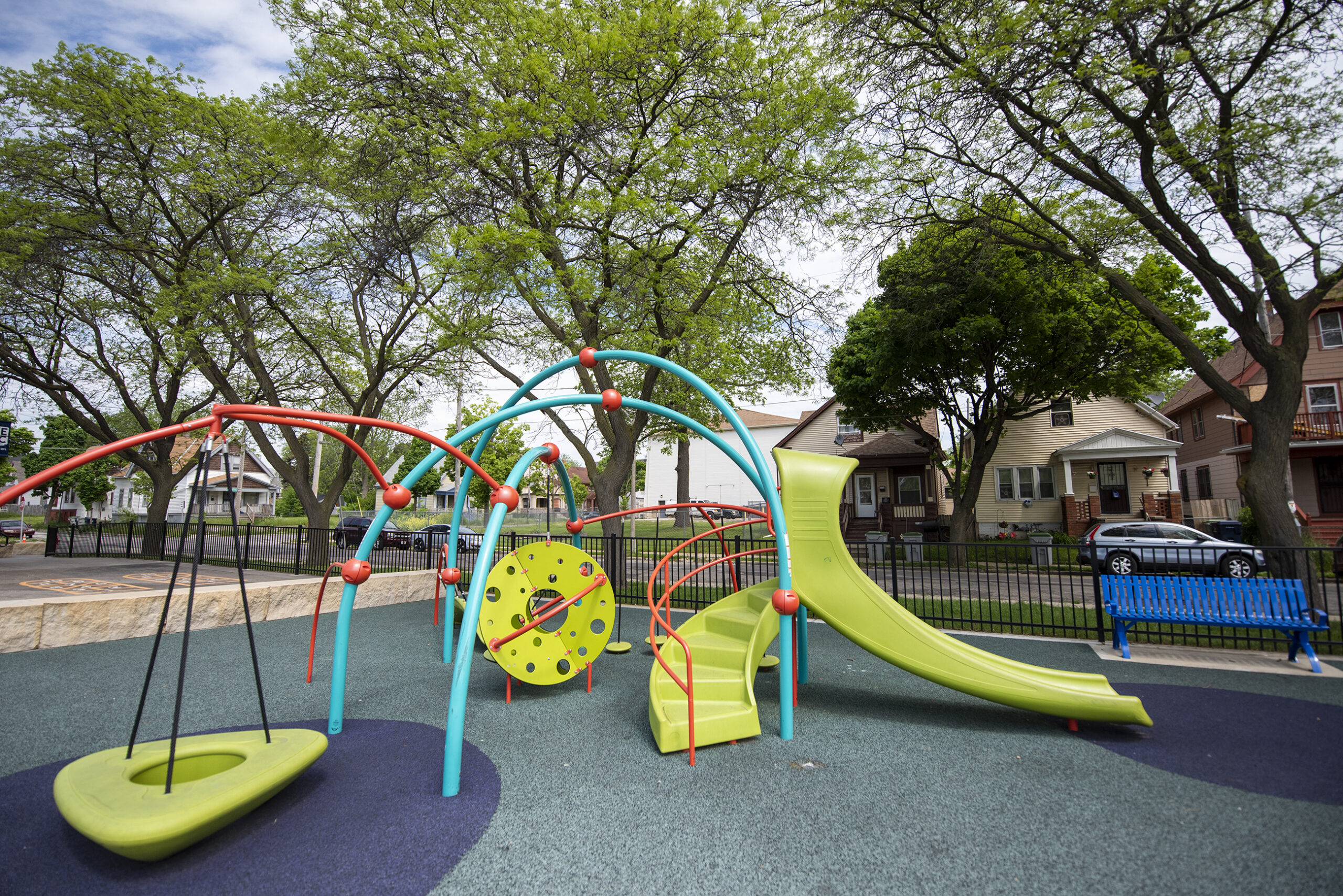 Green, blue and orange playground equipment is backdropped by green trees.