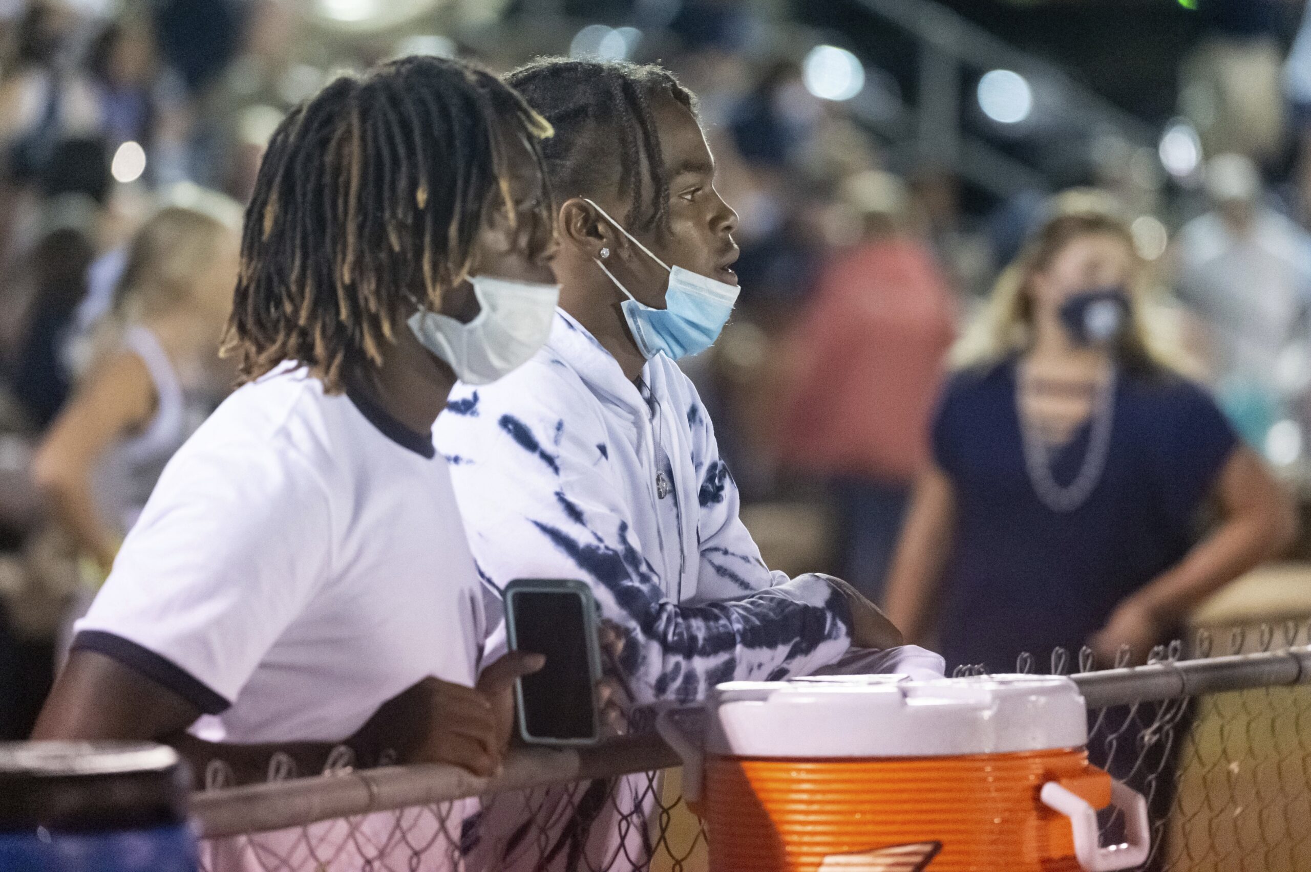 Two masked students watch high school football on sidelines