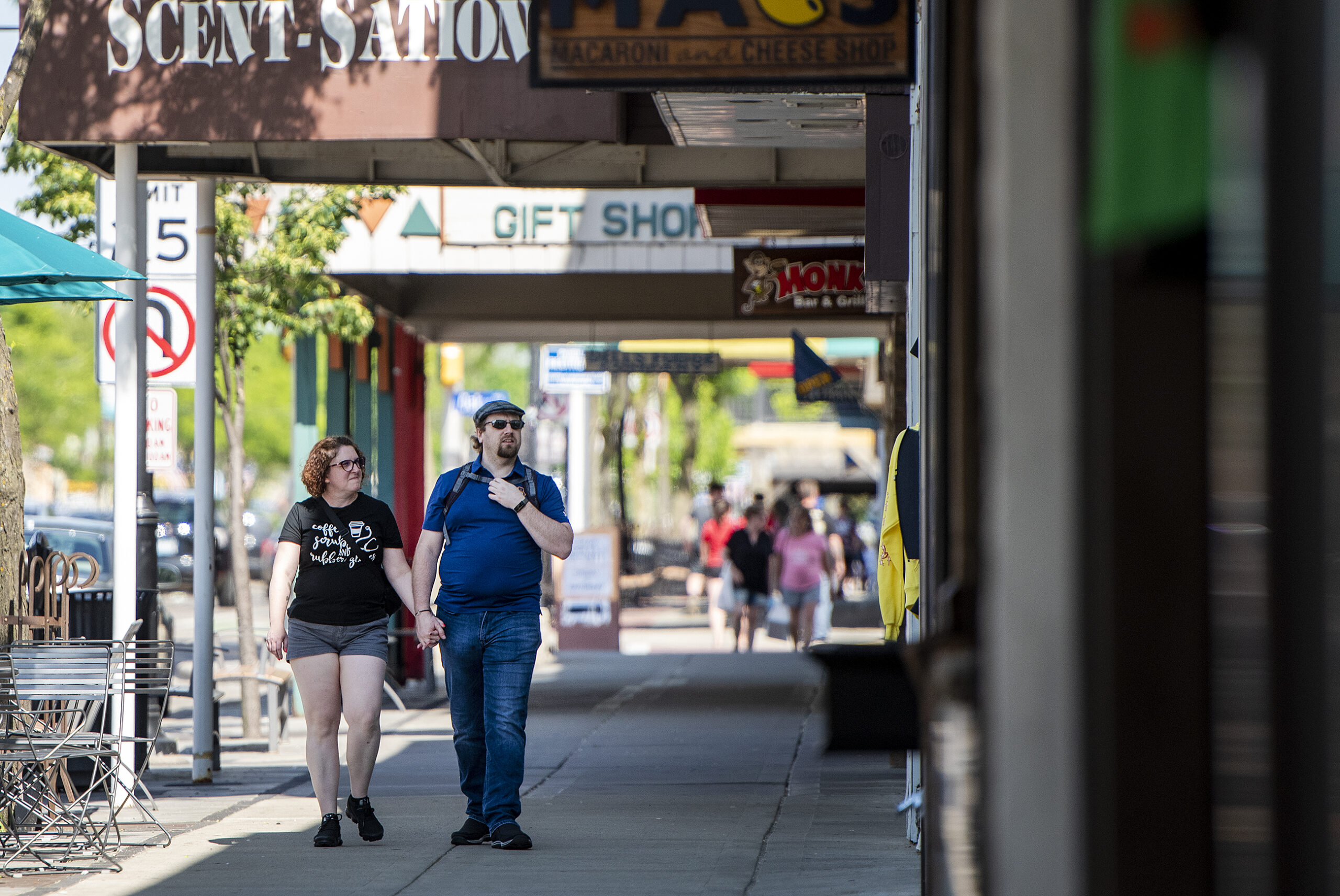 Two people walk on a sidewalk as they examine the exterior of stores on a sunny day.