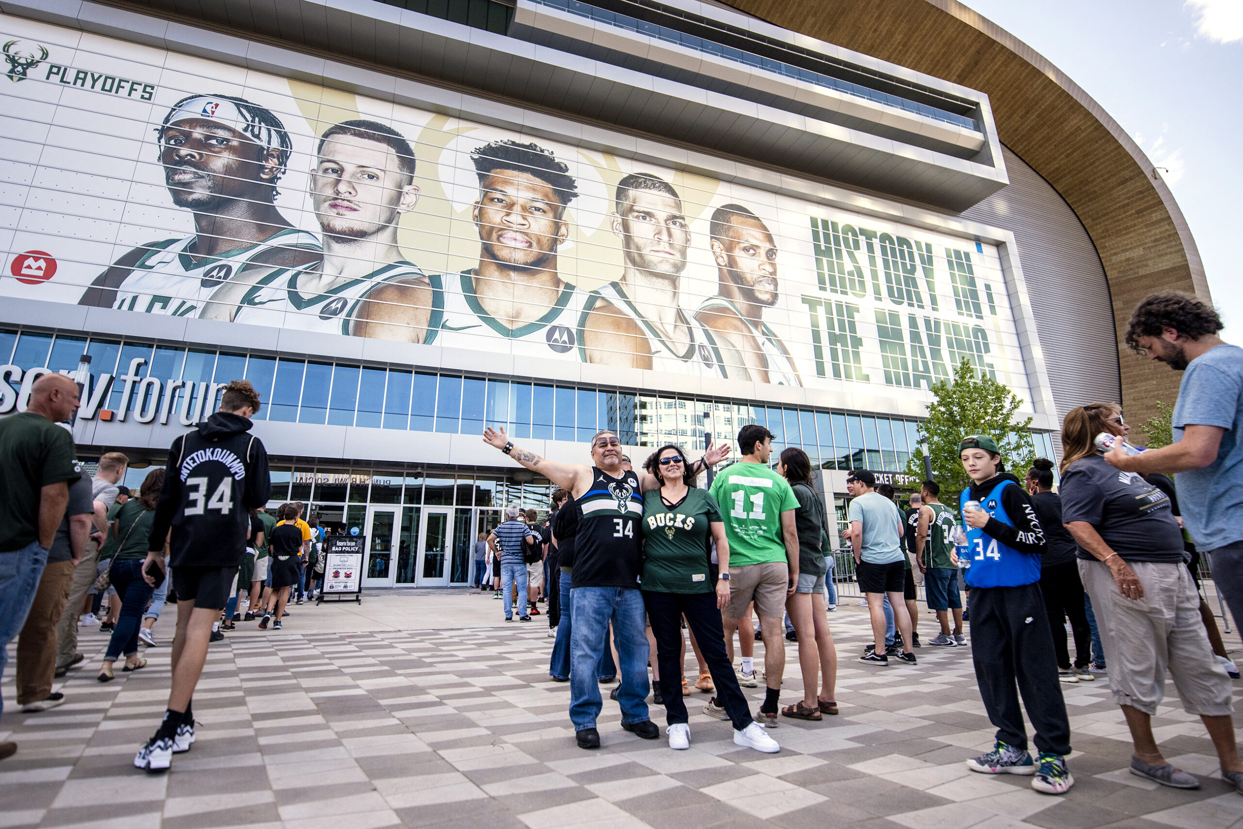 Fans raise their arms as they stand in front of the Fiserv Forum, which has large images of Milwaukee Bucks players on it.