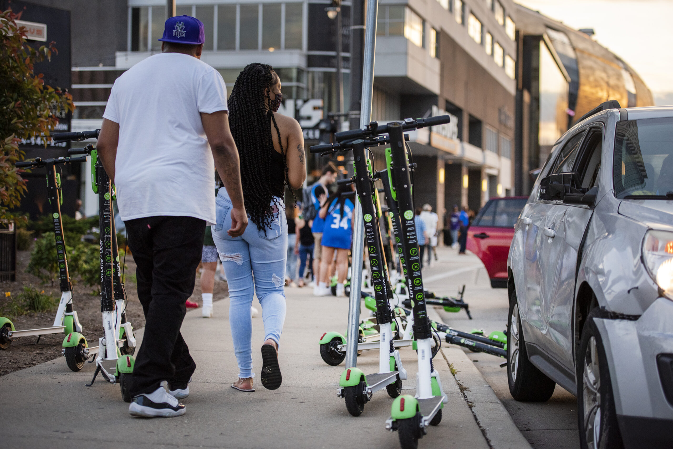 Many scooters are piled onto a sidewalk as pedestrians pass by.