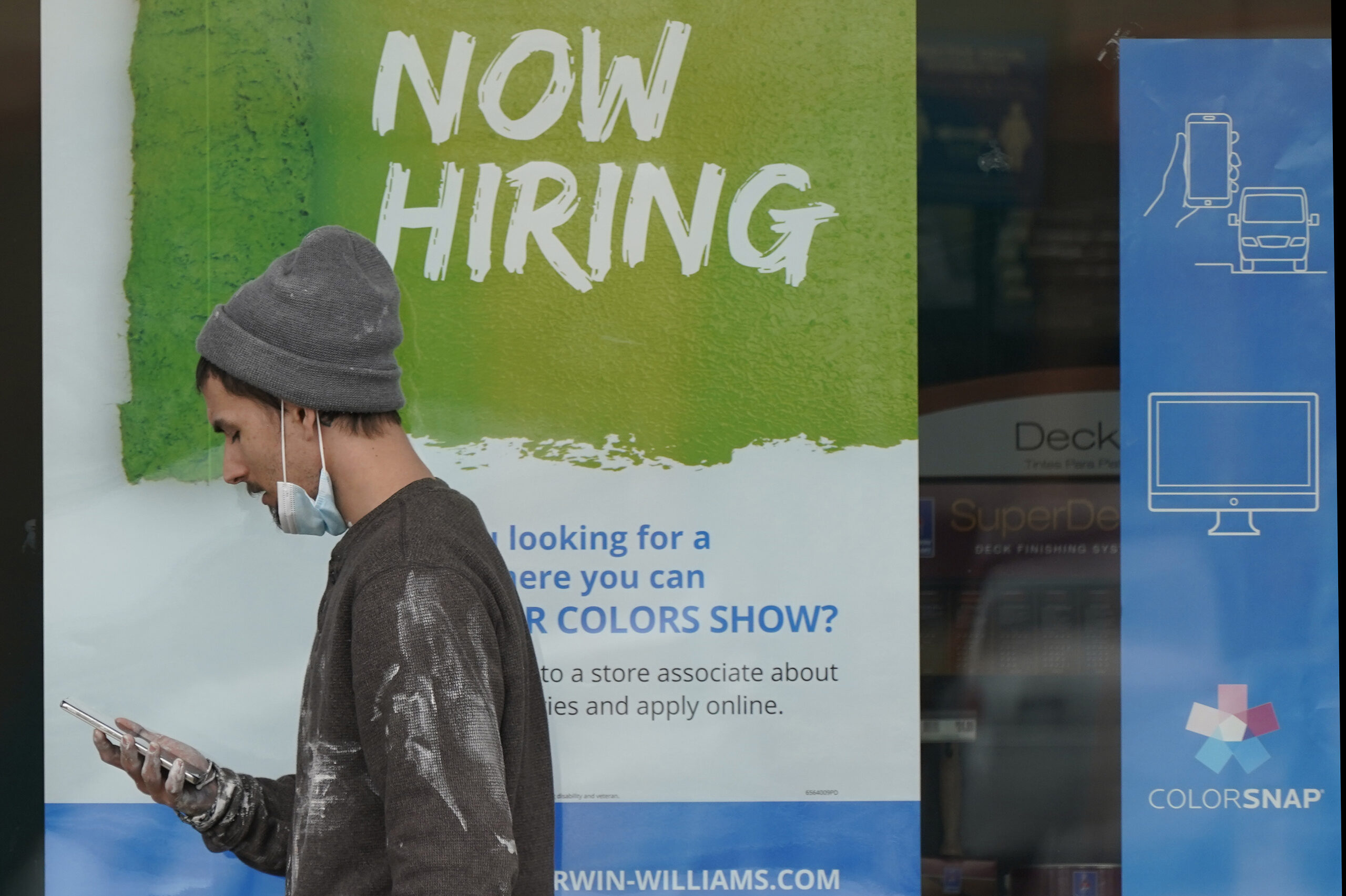 A man walks past a "Now Hiring" sign on a window at Sherwin Williams store