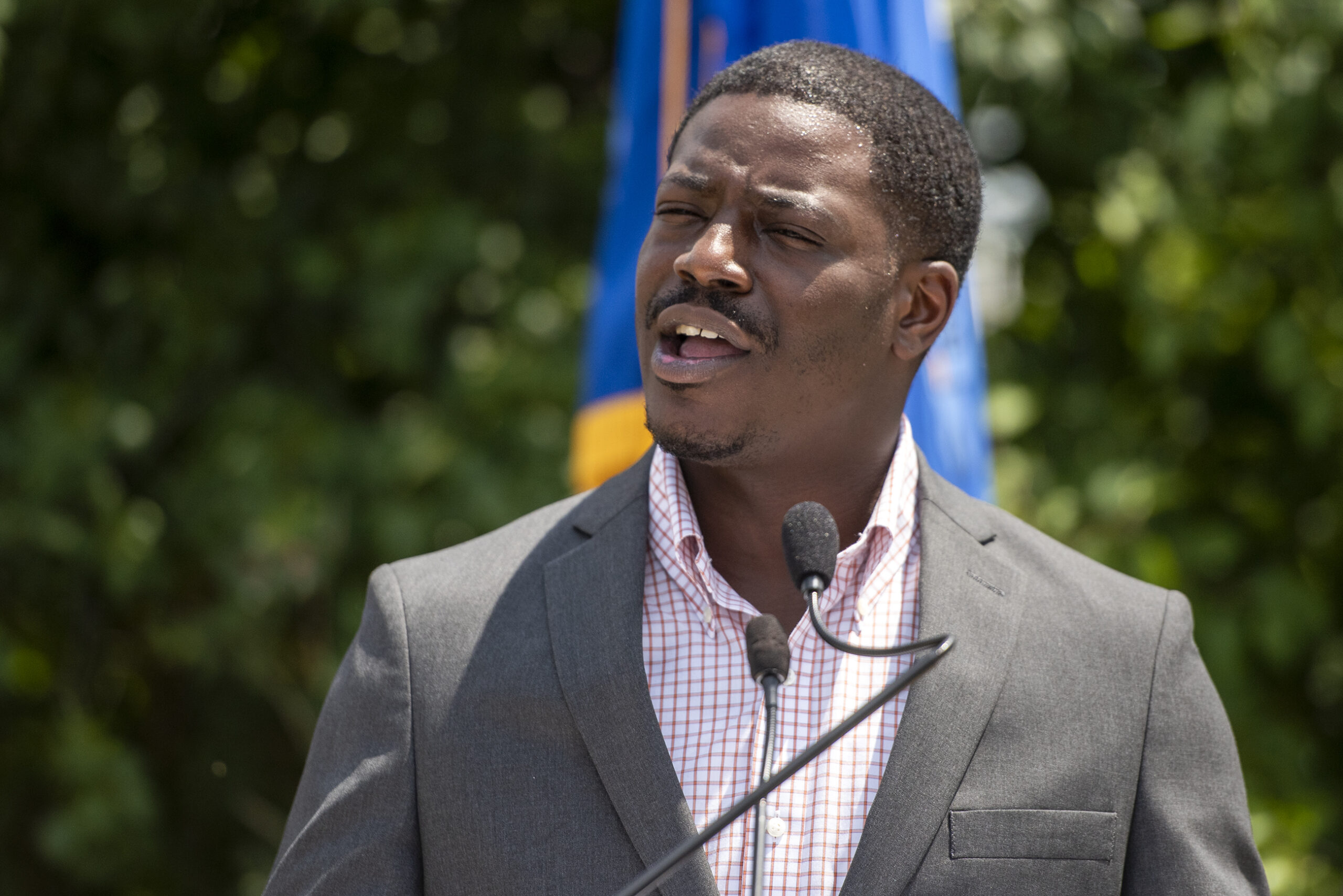 A man stands in front of green leaves as he speaks during an outdoor press conference.