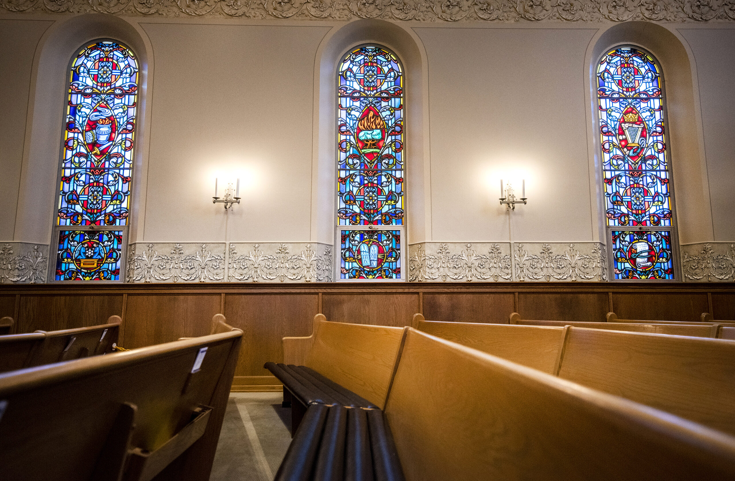 Three tall and narrow windows featuring vibrant stained glass are seen behind church pews.