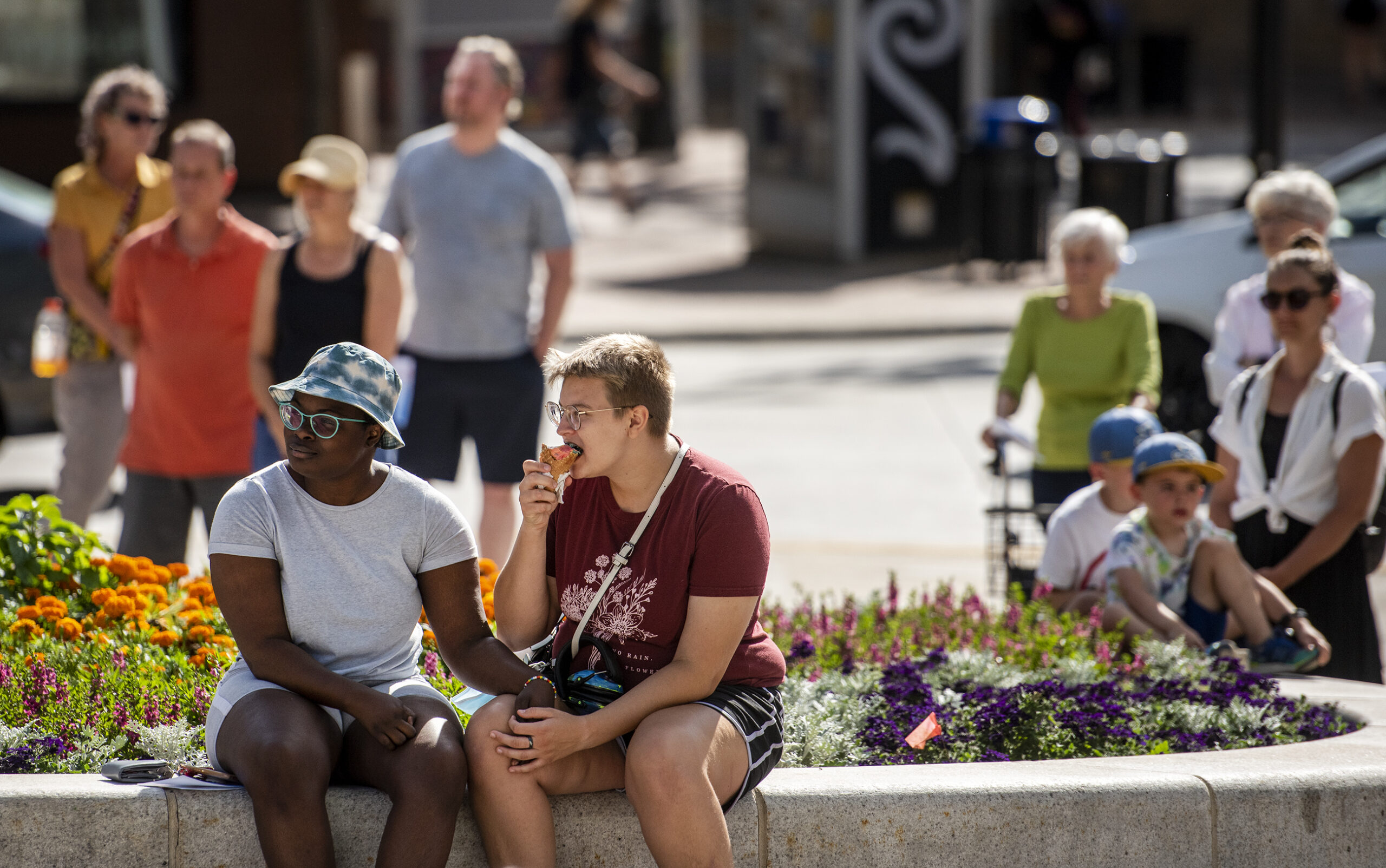 People sit and stand near flowers in on a sunny day in downtown Madison.