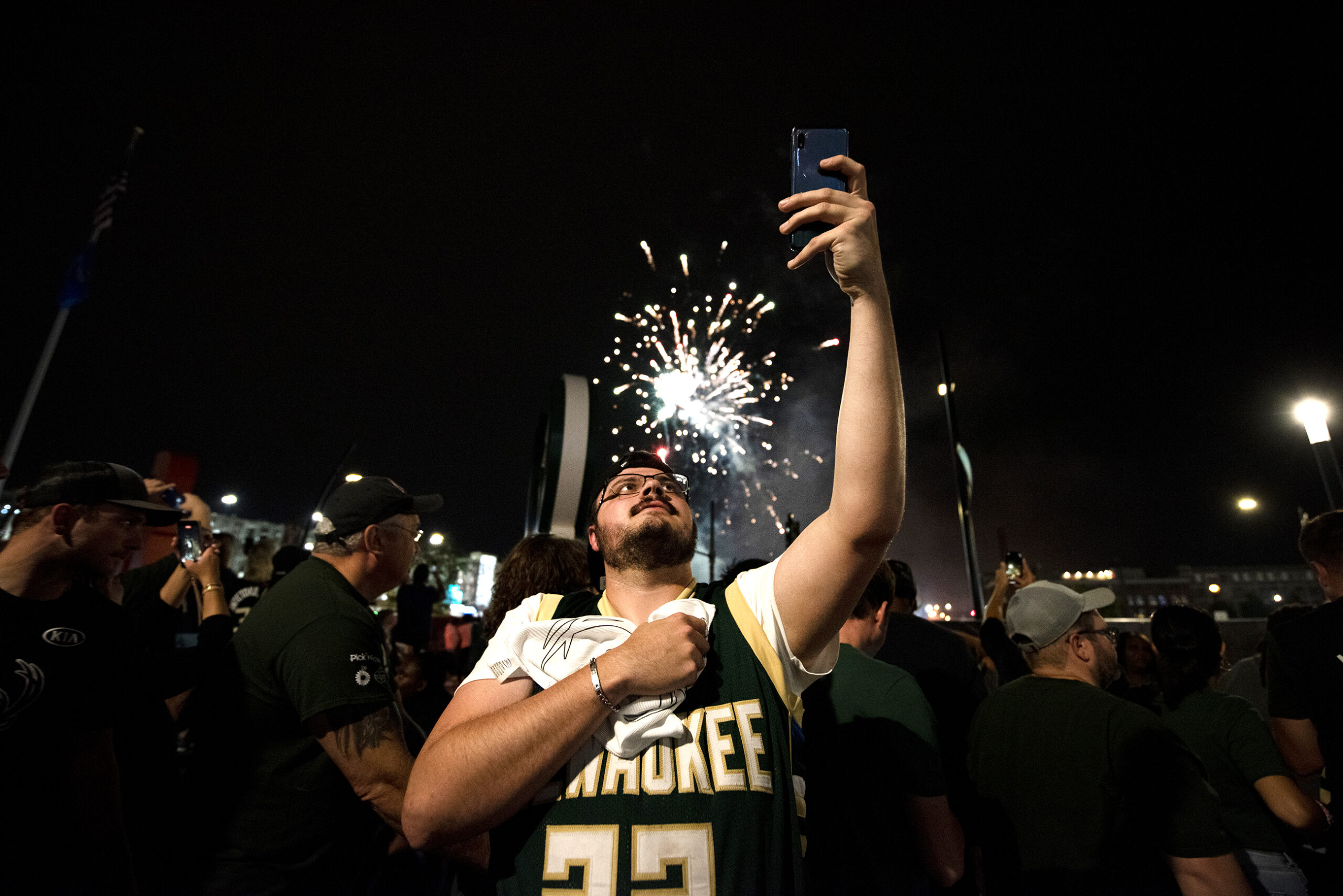 A fan in a Bucks jersey puts his hand over his heart as he records with his phone. Fireworks go off in the background.