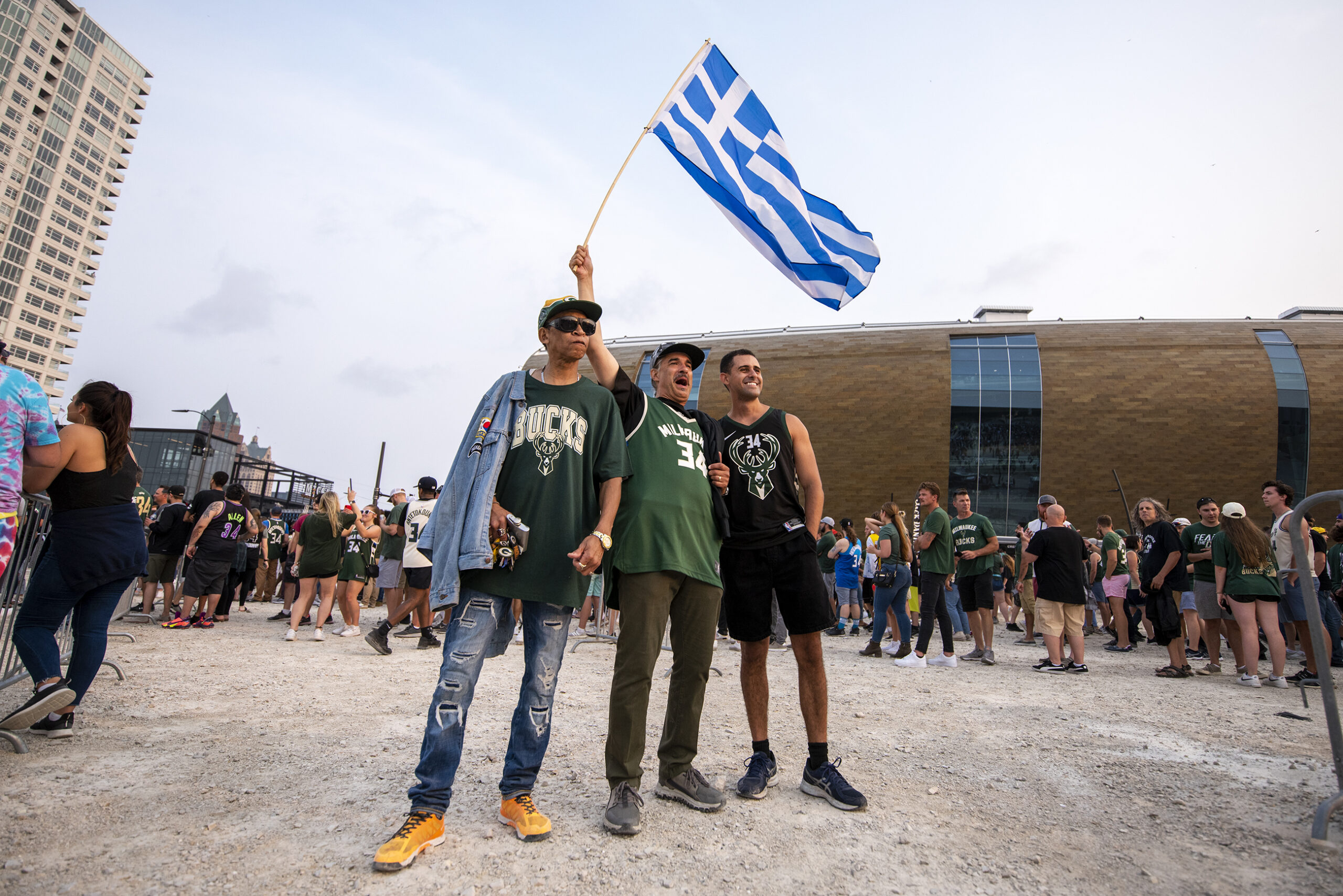 Three men stand together. One waves the blue and white Greek Flag up high.
