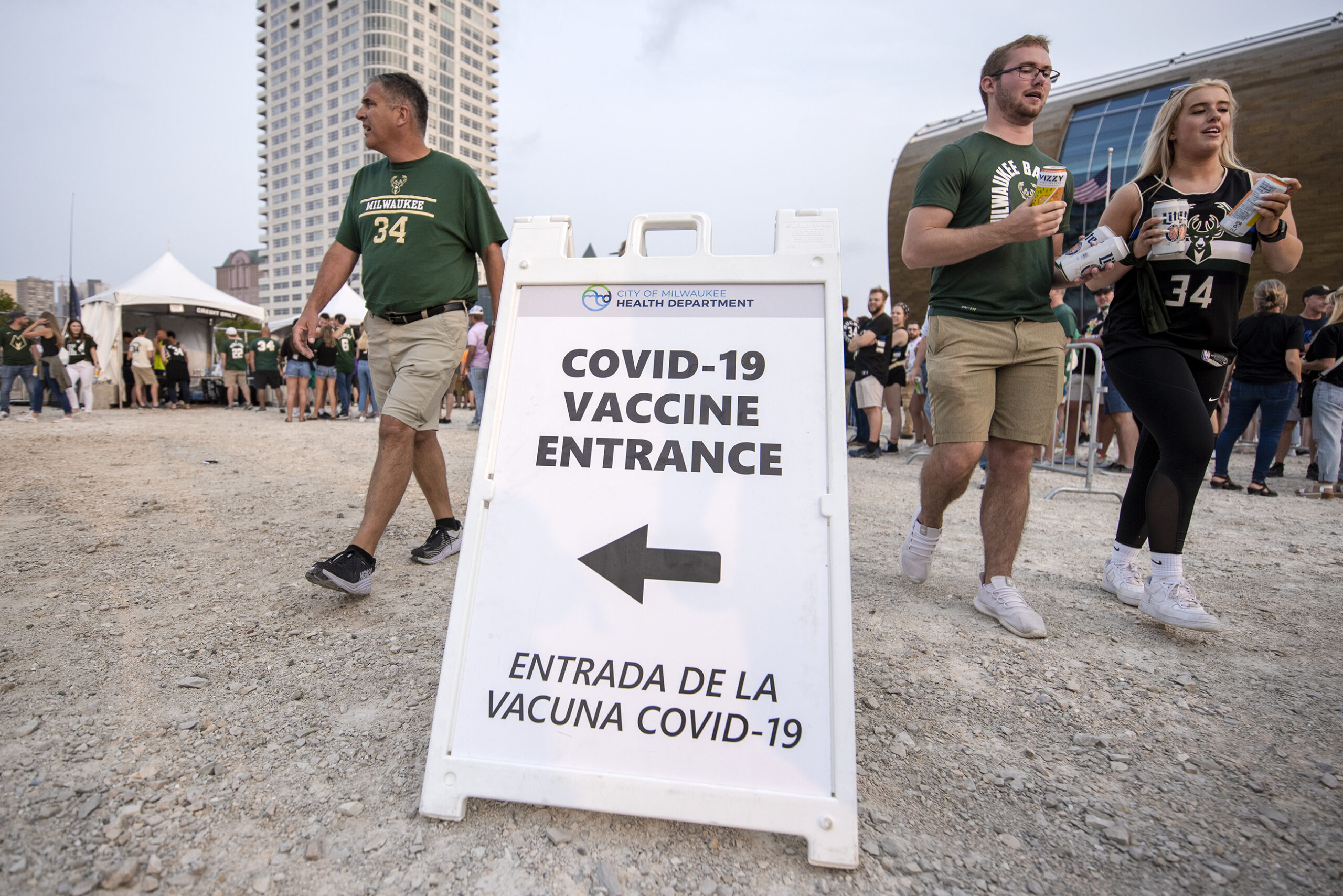 A sign says "COVID-19 Vaccine Entrance" as fans walk by holding drinks.
