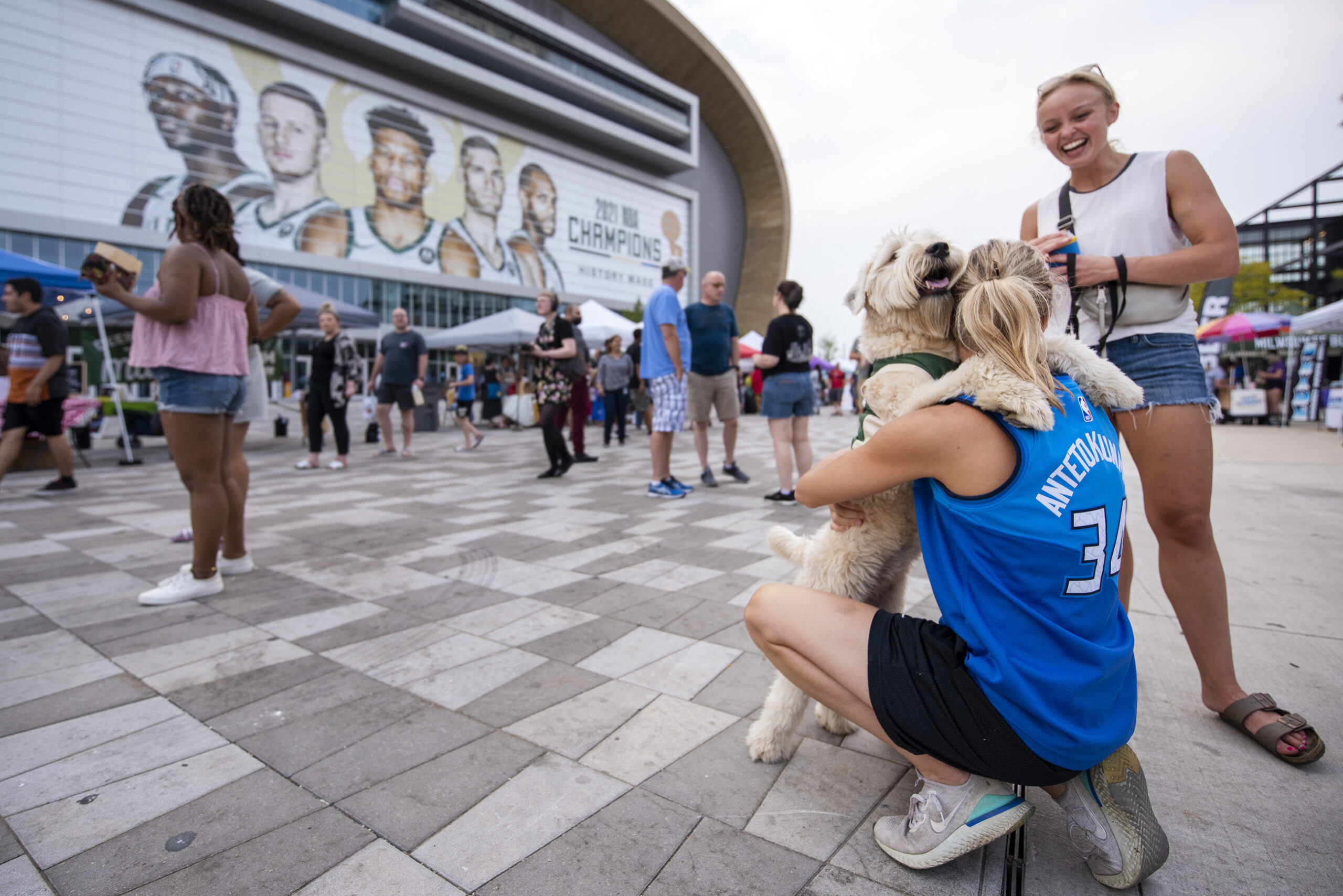 A woman wears a blue Bucks jersey as she hugs her dog with light fur in front of the Fiserv Forum.