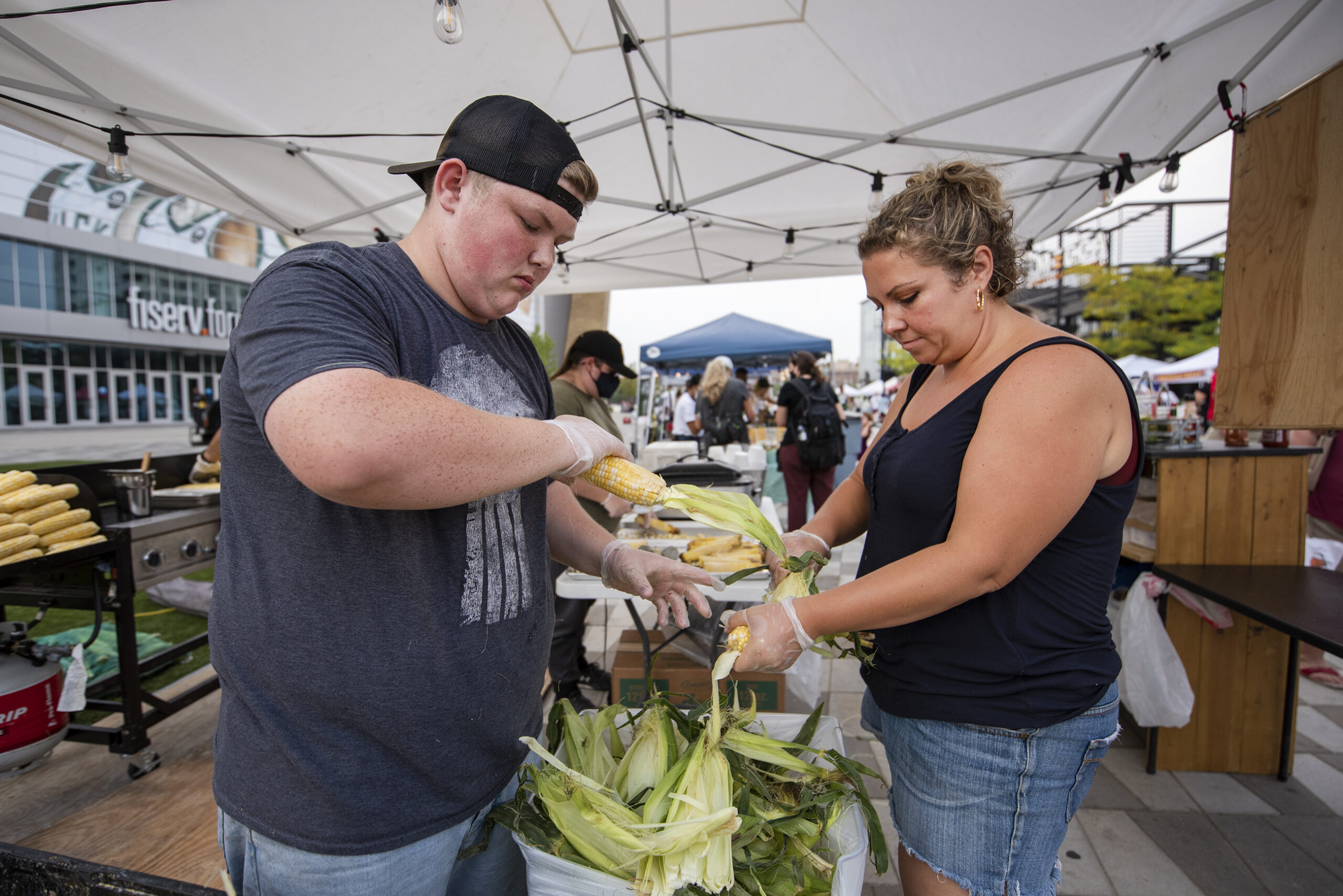 Two people work outdoors at a booth shucking corn.
