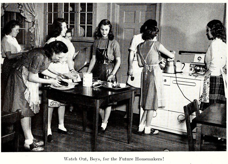 A black and white photo shows young women in aprons in the kitchen.