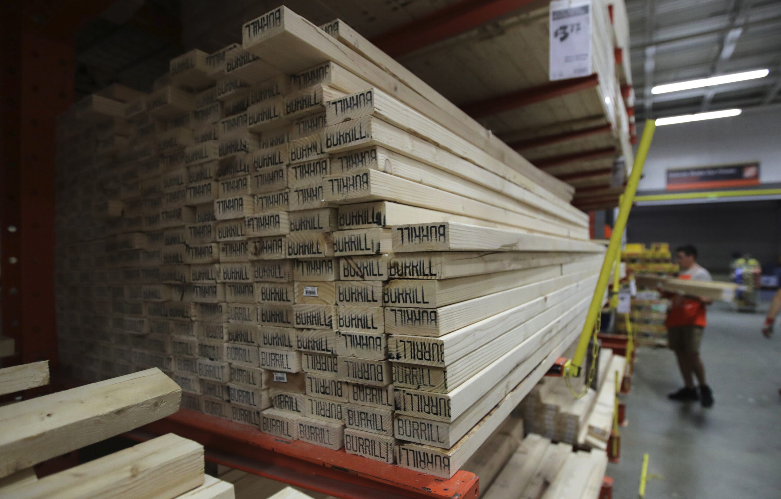 A pile of lumber sits in a Home Depot with an employee in the background