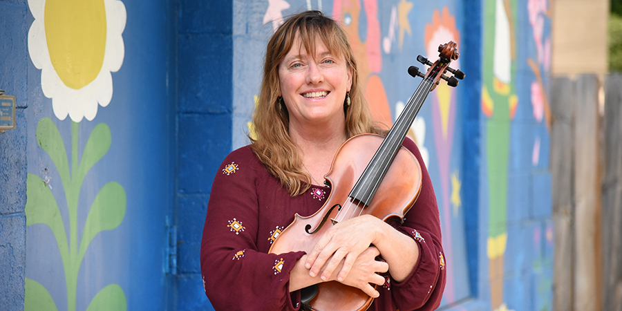 Sile Shigley holds a fiddle standing in front of a colorful mural wall