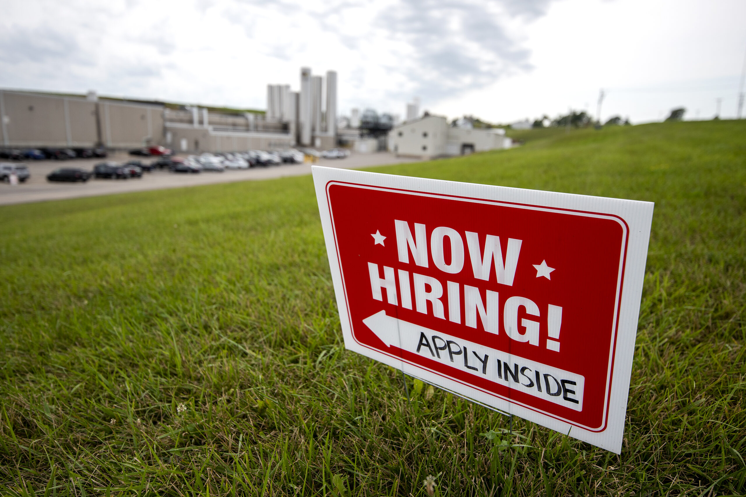 A red "Now Hiring!" sign located in a grassy field near a county road instructs potential applicants to apply outside.